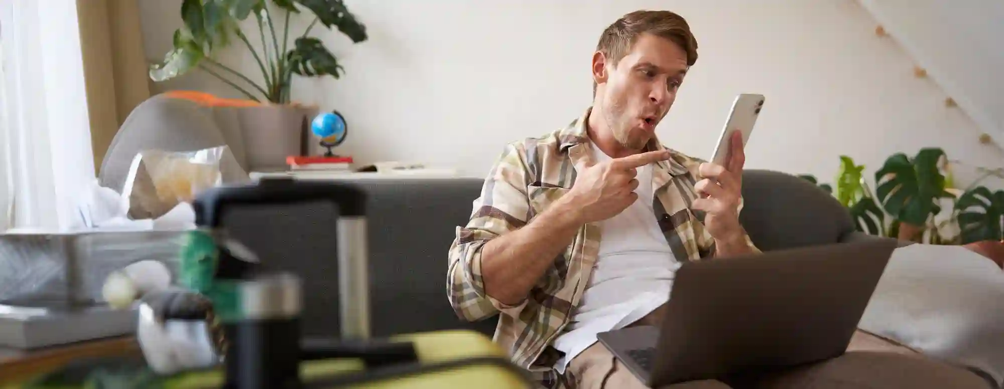 Man on couch points excitedly at his phone while packing for a trip, using a laptop.