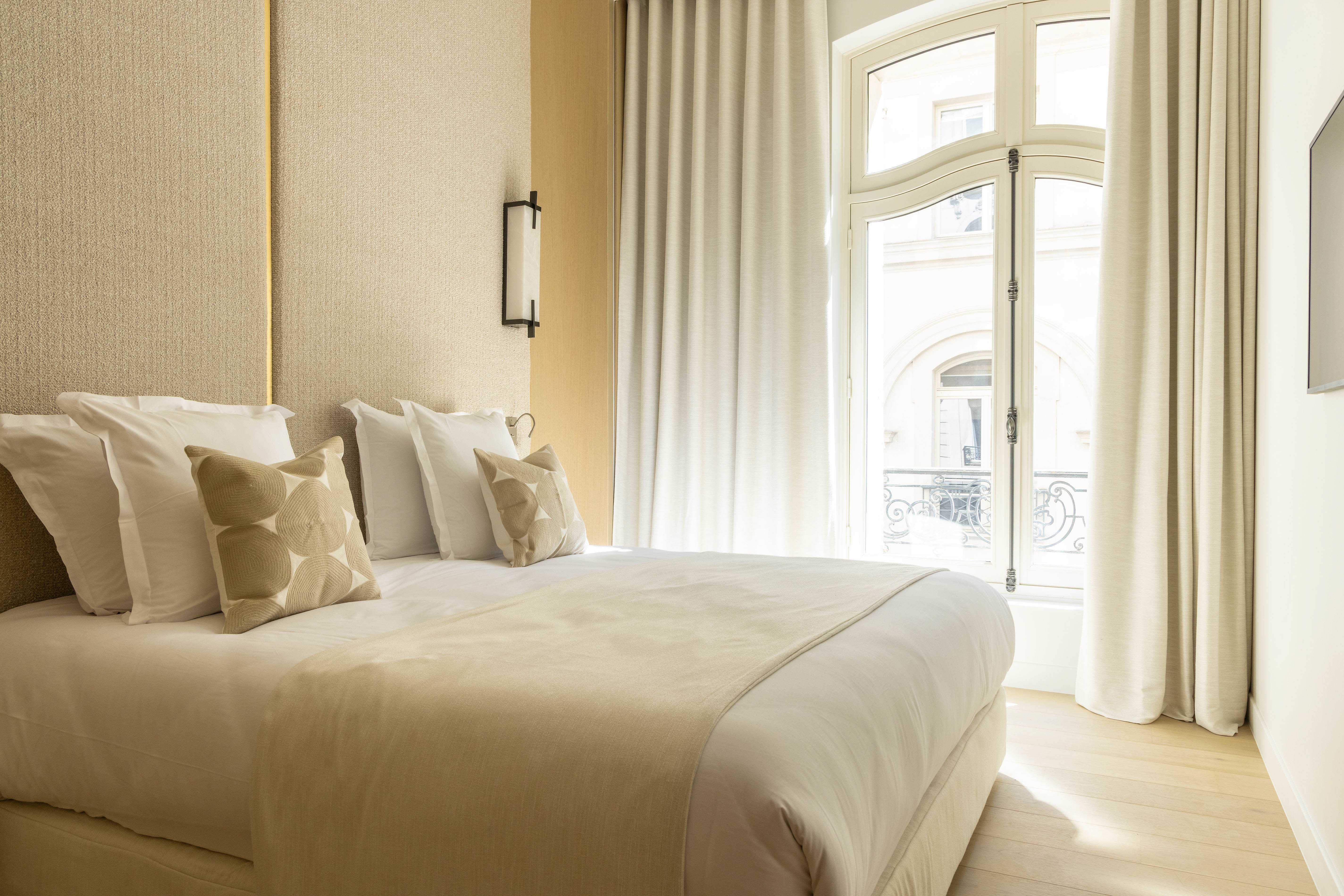 Chambre seconde avec lit double, tete de lit beige, coussins dores, grande fenetre cintree et lumiere naturelle