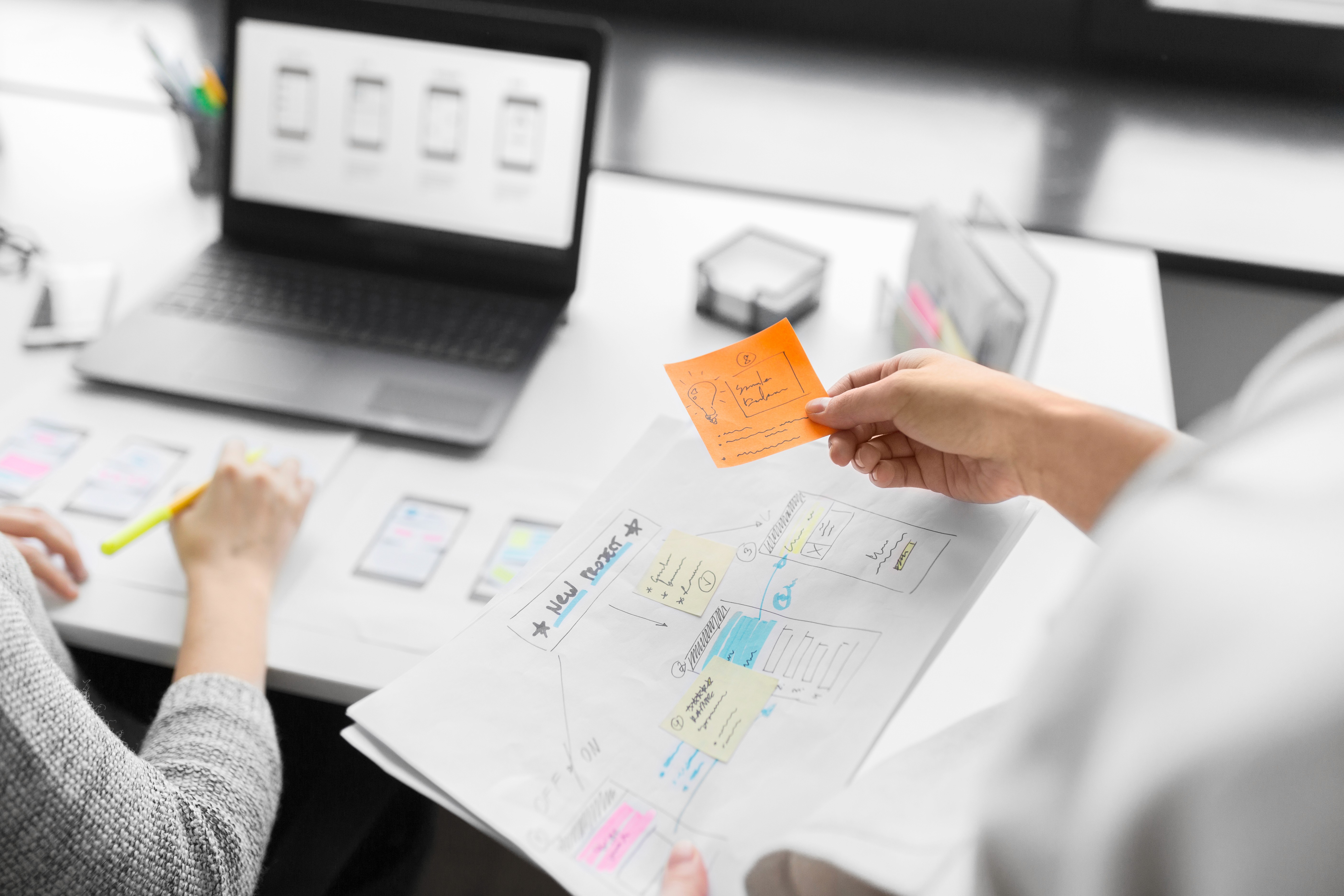 Hand hält orangefarbenen Notizzettel über einem Papier mit Skizzen für Benutzeroberflächen, im Hintergrund Laptop und weitere Papiere auf einem Tisch