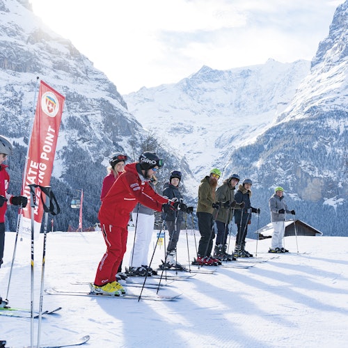 一群穿着滑雪装备的滑雪者在雪地上排成一排，背景是群山，旁边竖立着一个“集合点”标志。