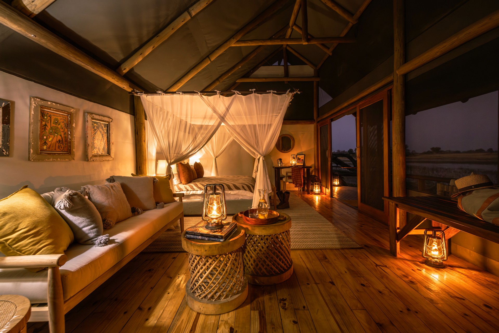 Kujwana Delta Ride, Okavango Delta, Botswana – Riverside Suites på Kujwana Camp, lyxigt boende med panoramavy över Okavango i kvällsljus. Vackert sken från lyktor och lampor belyser den stora dubbelsängen, vackert bäddad och omgiven av myggnät.