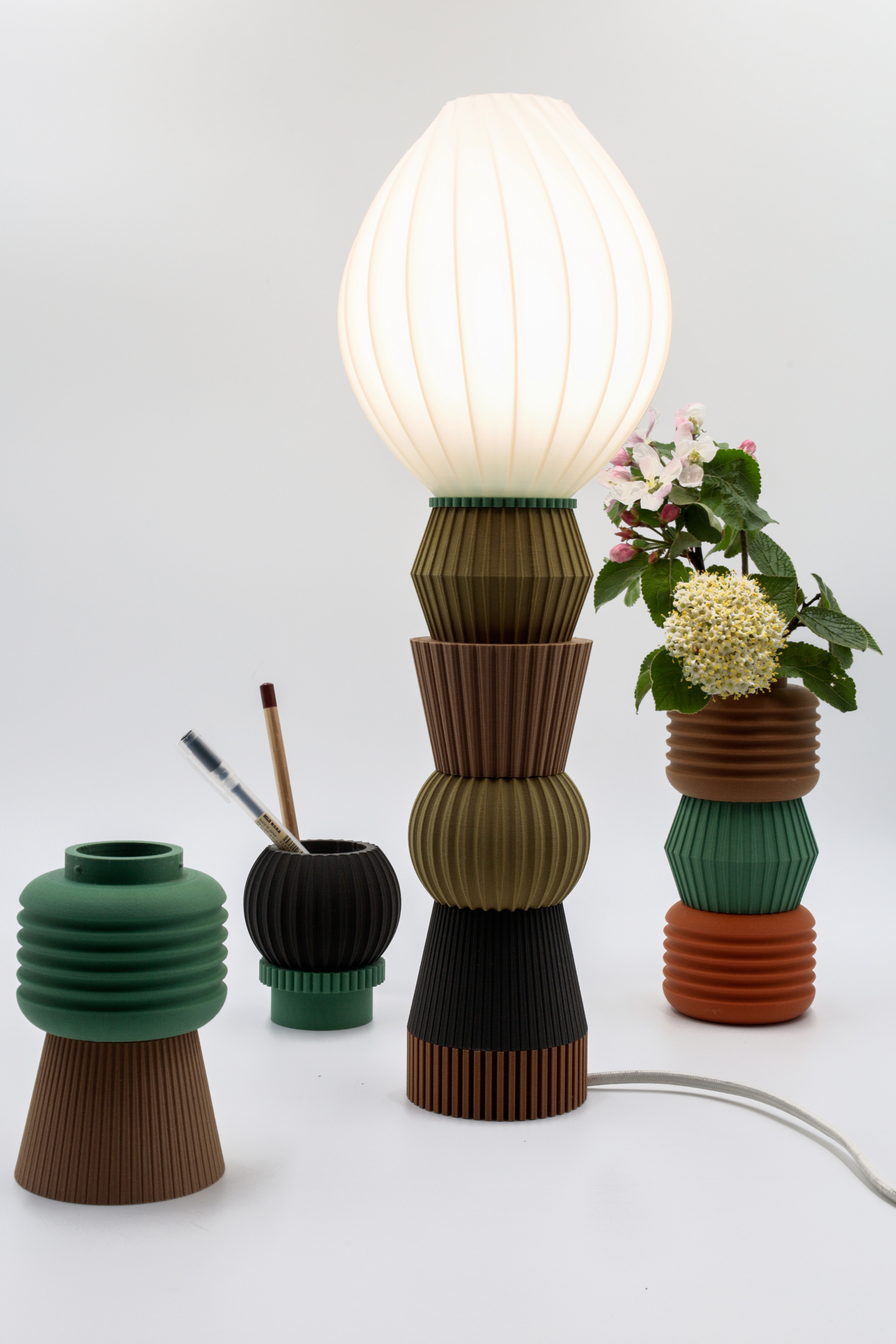 An array of artistic lamp bases composed of stacked, colorful, ribbed elements, with one featuring an illuminated lampshade on top, and others creatively repurposed to hold writing brushes and a bouquet of flowers.