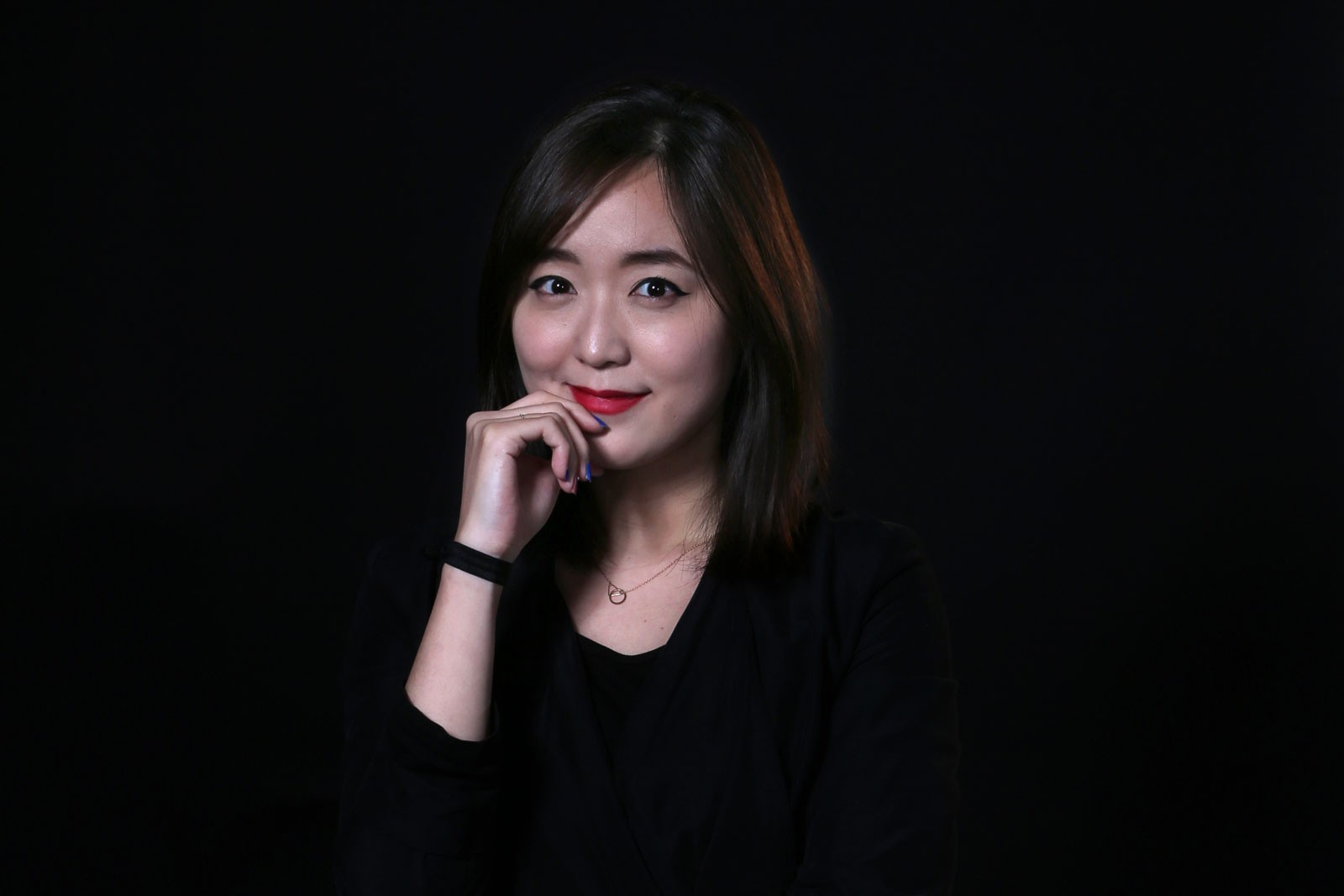 A young woman with shoulder-length dark hair and light skin is shown against a solid black background. She is wearing a black top and a thin silver necklace. Her right hand is touching her chin, and her left hand is partially visible near her wrist. She is looking directly at the camera with a slight smile.