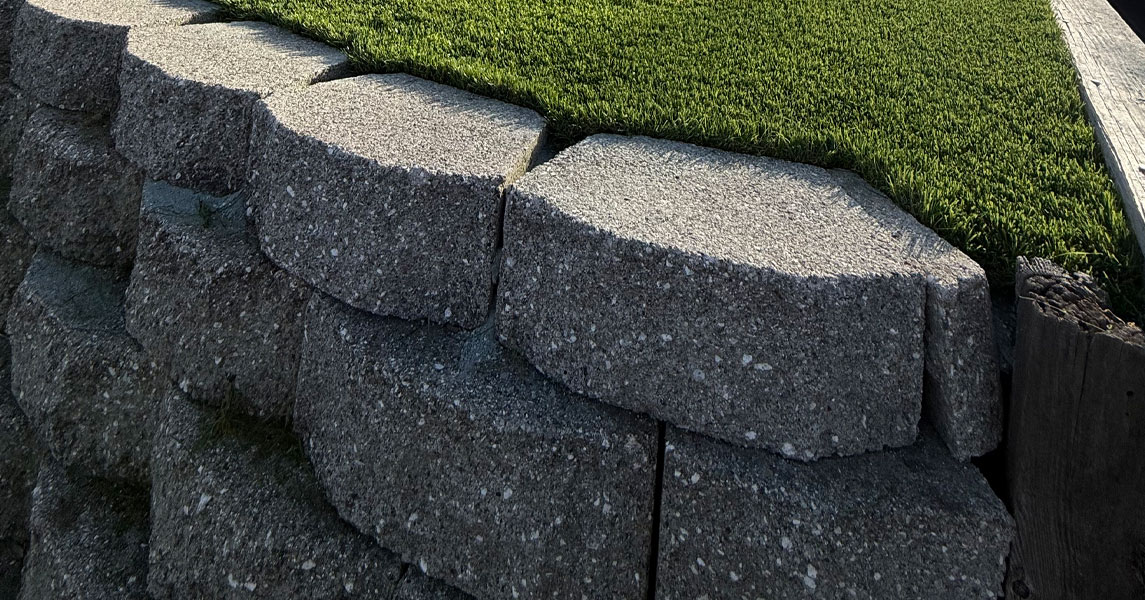 Stacked-block retaining wall in Hercules shown close up, with capstones meeting an artificial turf lawn edge.