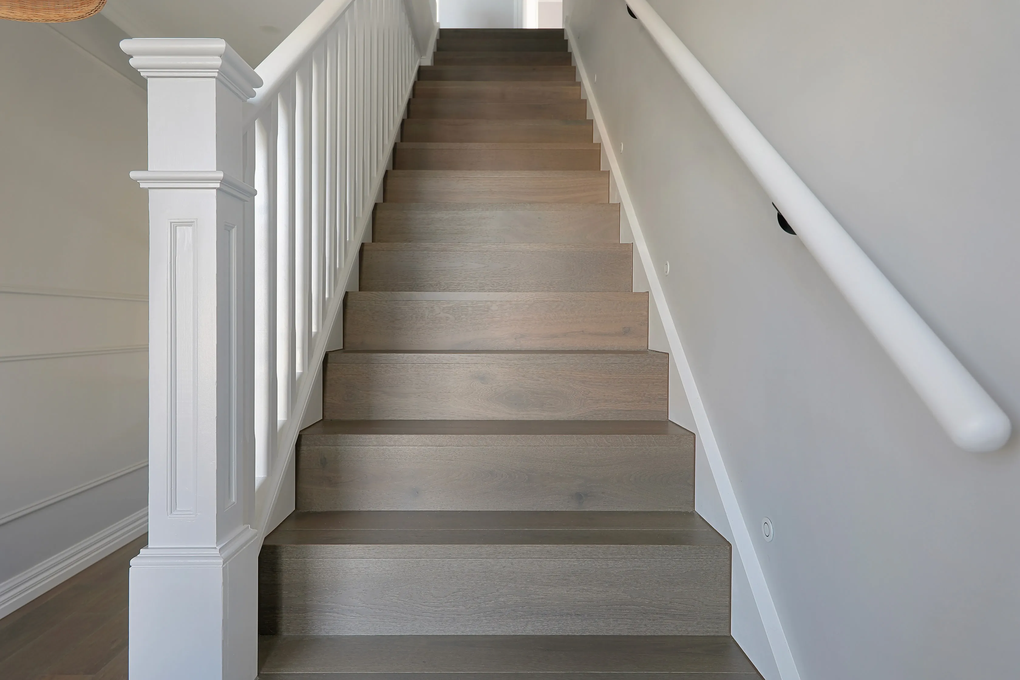 Hamptons staircase with white panelled newel posts and balustrade, a white wall handrail and warm grey-oak treads against panelled walls