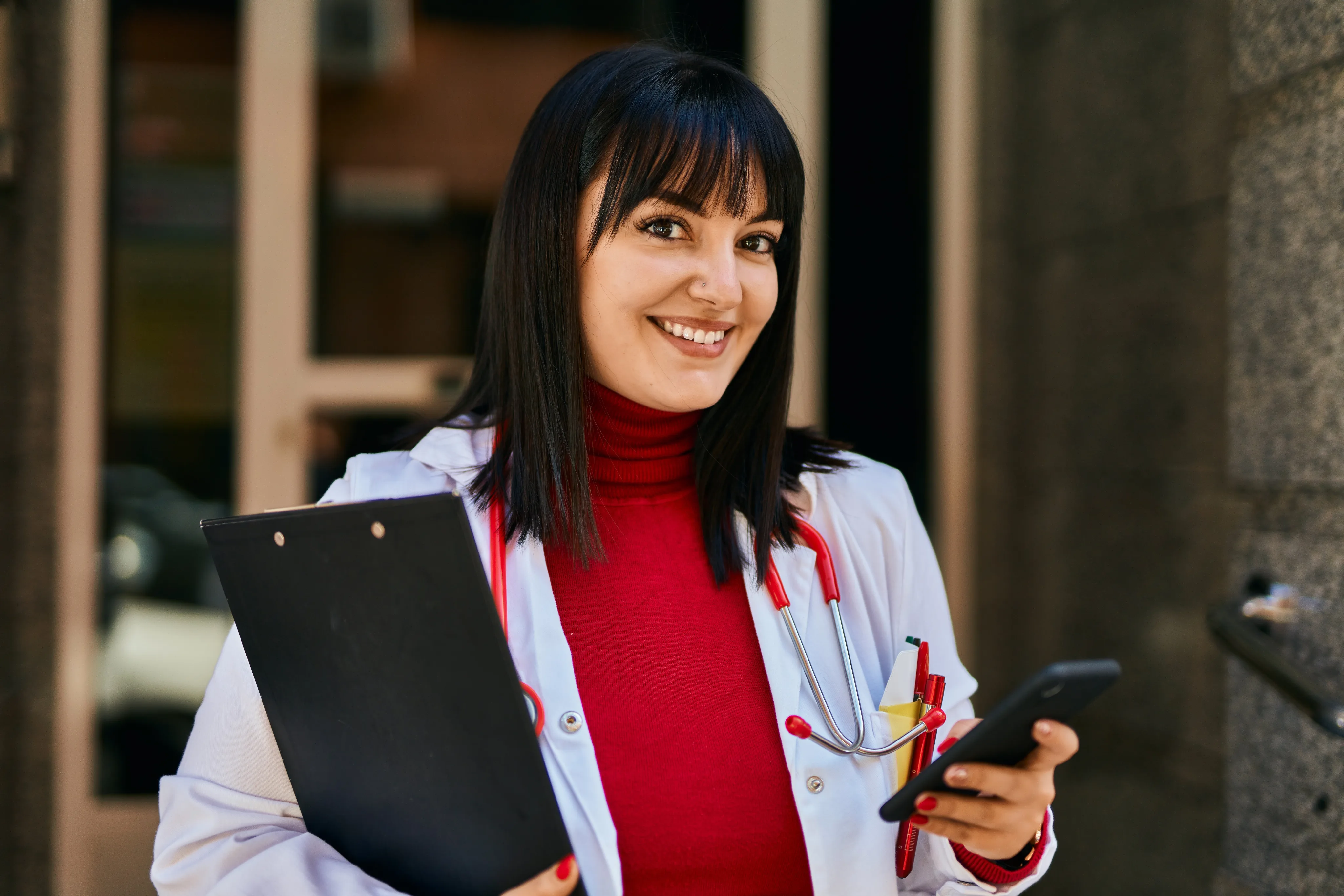 Smiling female doctor with dark hair and bangs wearing a white coat and red turtleneck, holding a clipboard and smartphone in a modern medical facility.