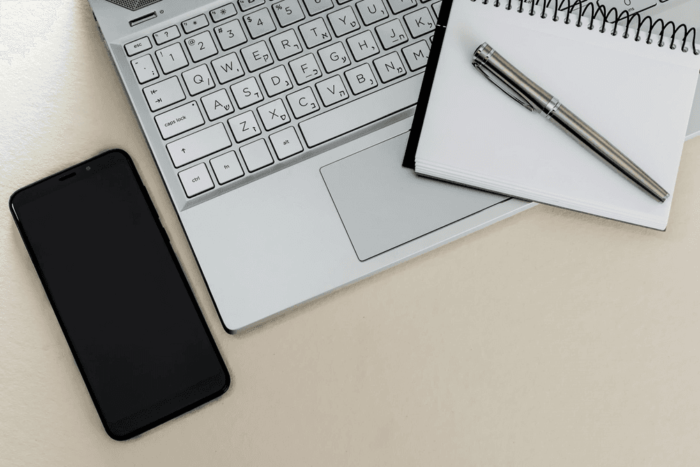Laptop keyboard with a notebook, pen, and smartphone on a desk.