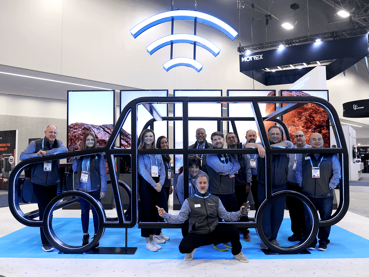 A group of people pose and smile inside a large, stylized car frame display at an indoor event. Blue Wi-Fi symbol lights hang above the display, and screens show rocky landscapes in the background.