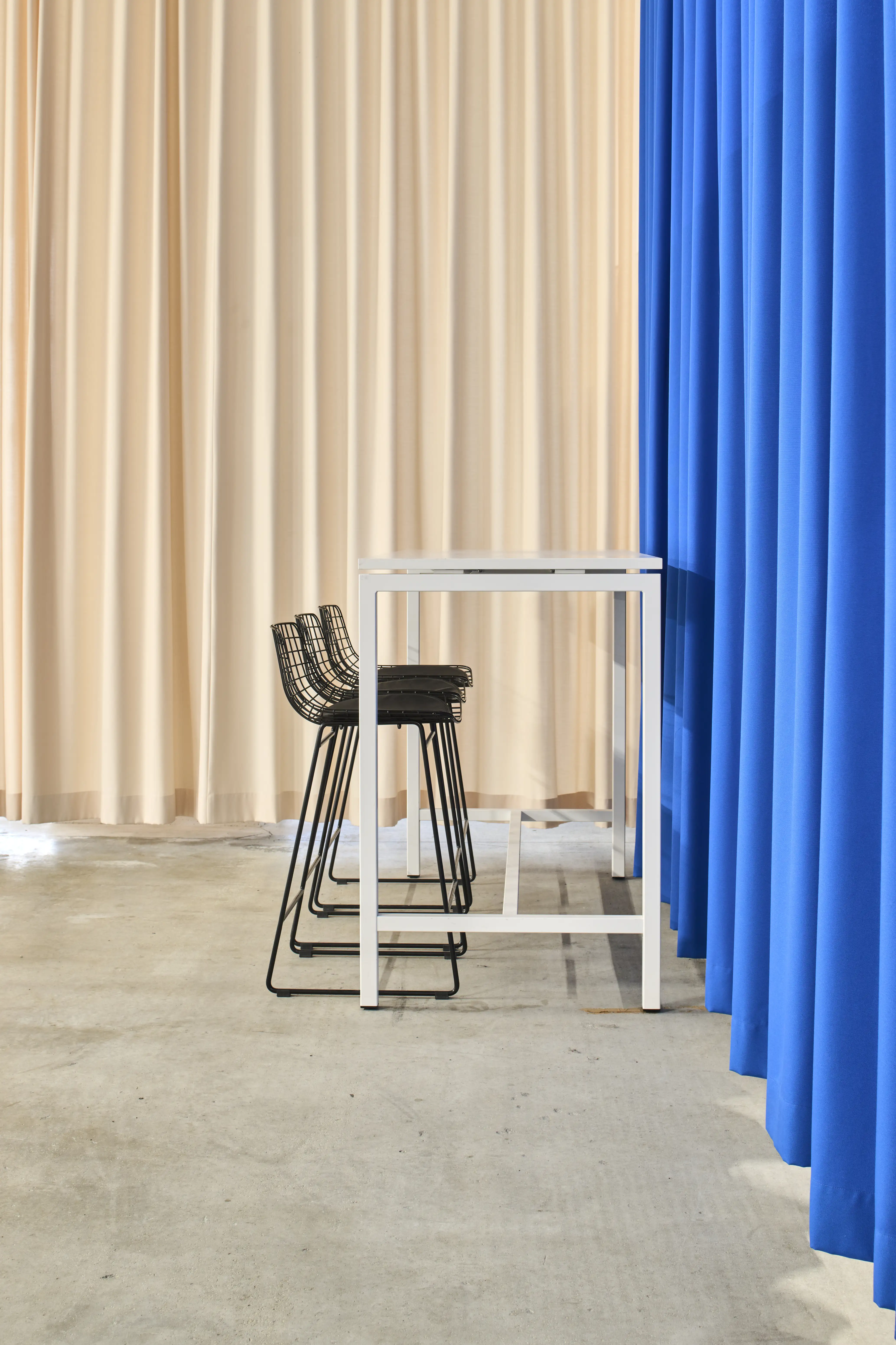 White high table with black wire bar stools at the FLACONI CONTENT STUDIO, framed by a beige and a bright blue floor-length curtain.