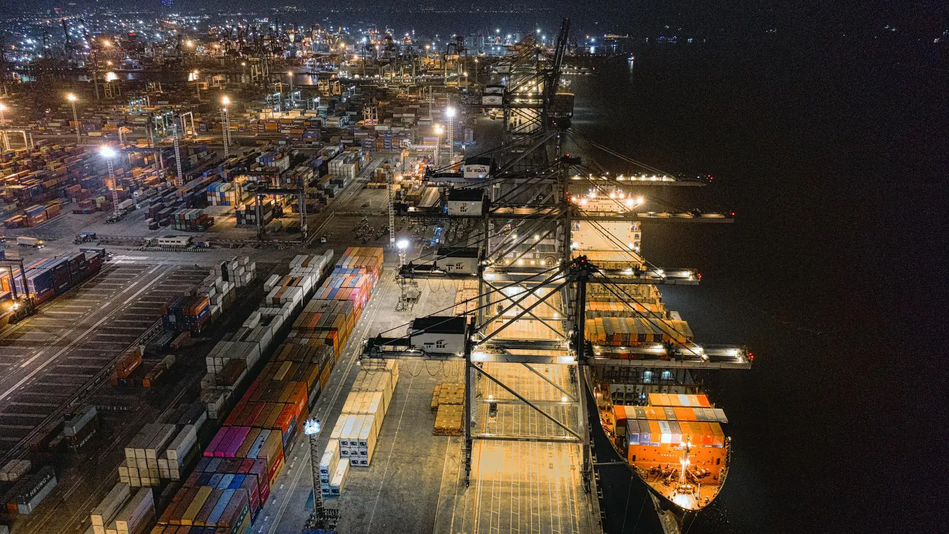 Aerial view of a bustling port at night, illuminated by bright lights. Cranes loom over colorful shipping containers and a docked cargo ship, conveying busy industrial activity.