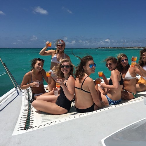 A group of women in swimsuits enjoy drinks on a boat, with turquoise ocean and blue sky in the background.