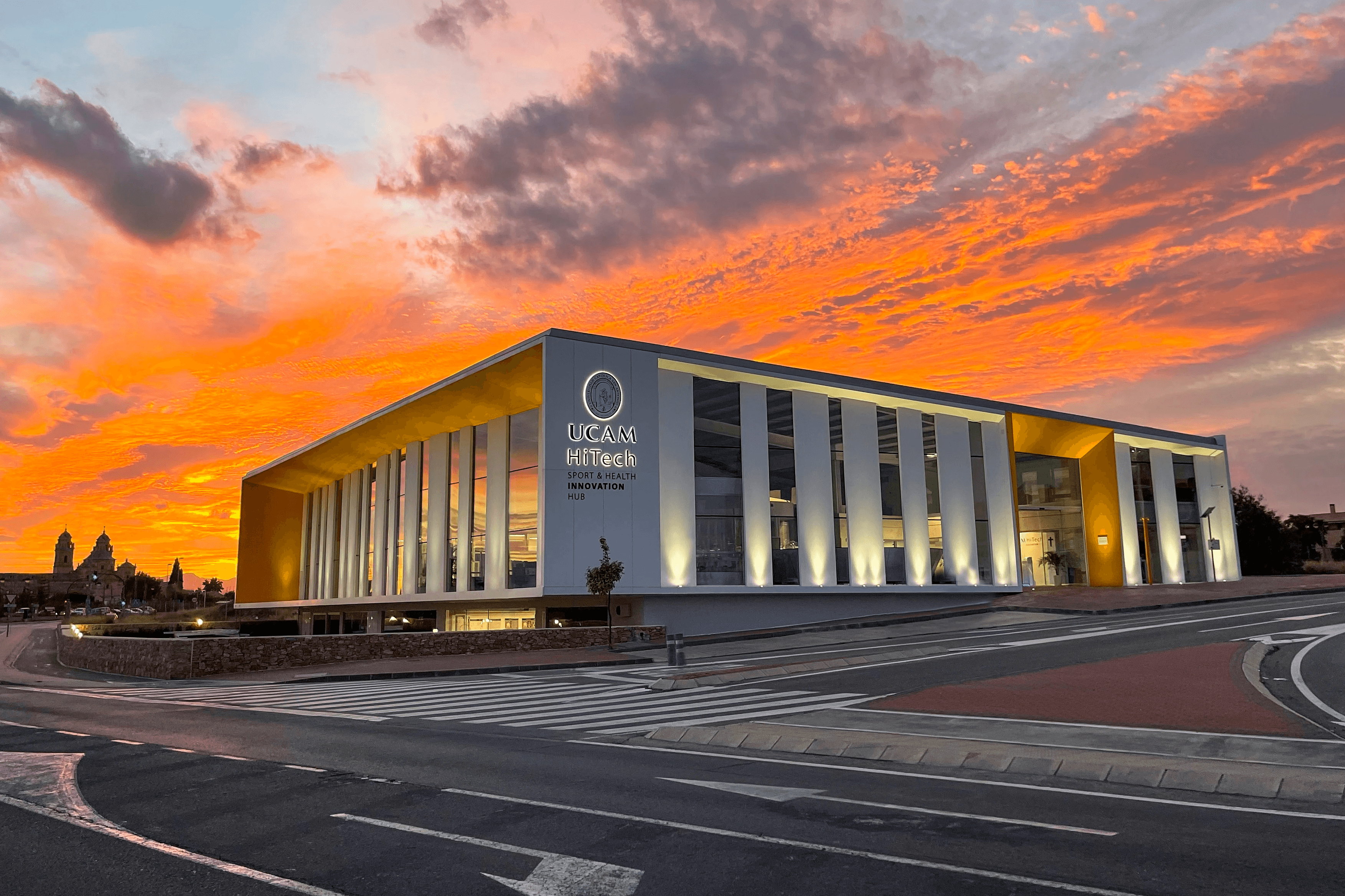 Three people are enjoying a meal at an outdoor café near a historic building, surrounded by lush green trees, with one person wearing a shirt featuring European UCAM University.