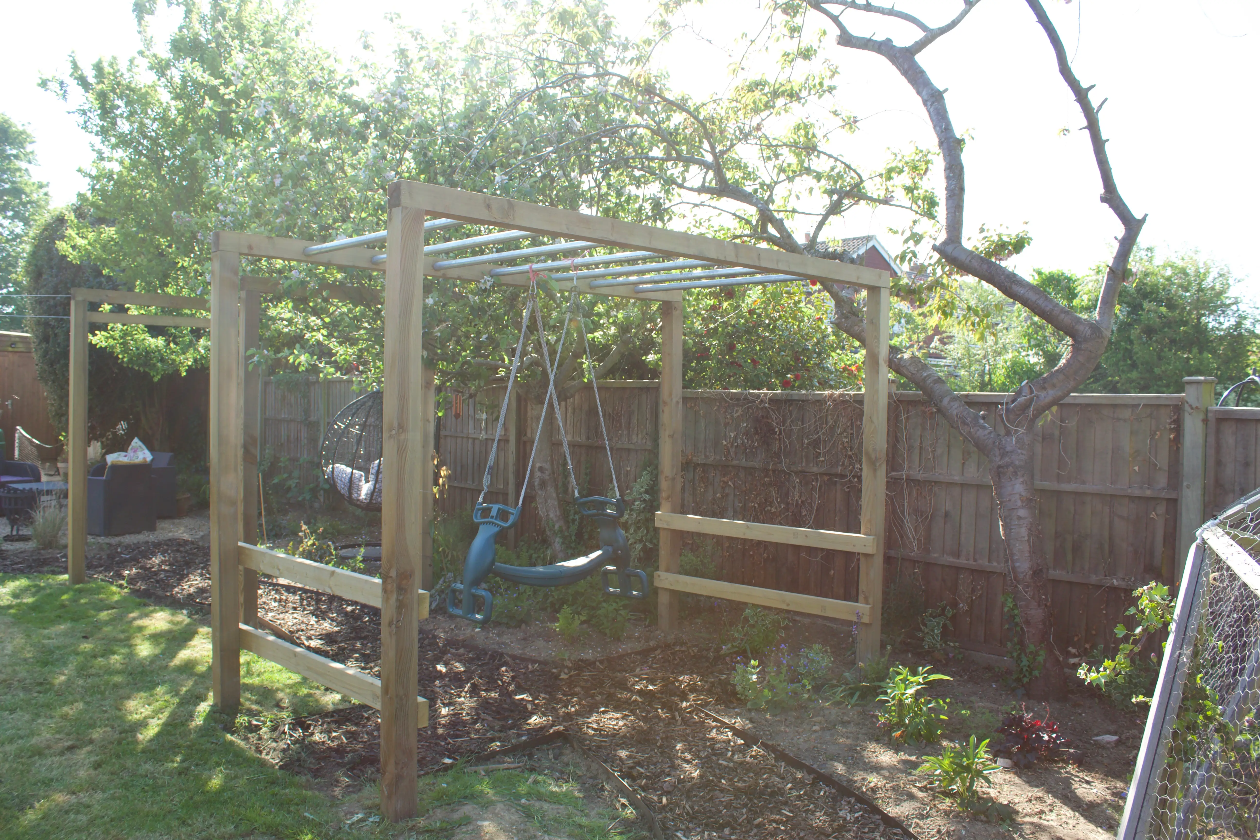 A wooden structure surrounded by greenery, partially shaded by trees, with a rustic fence in the background.