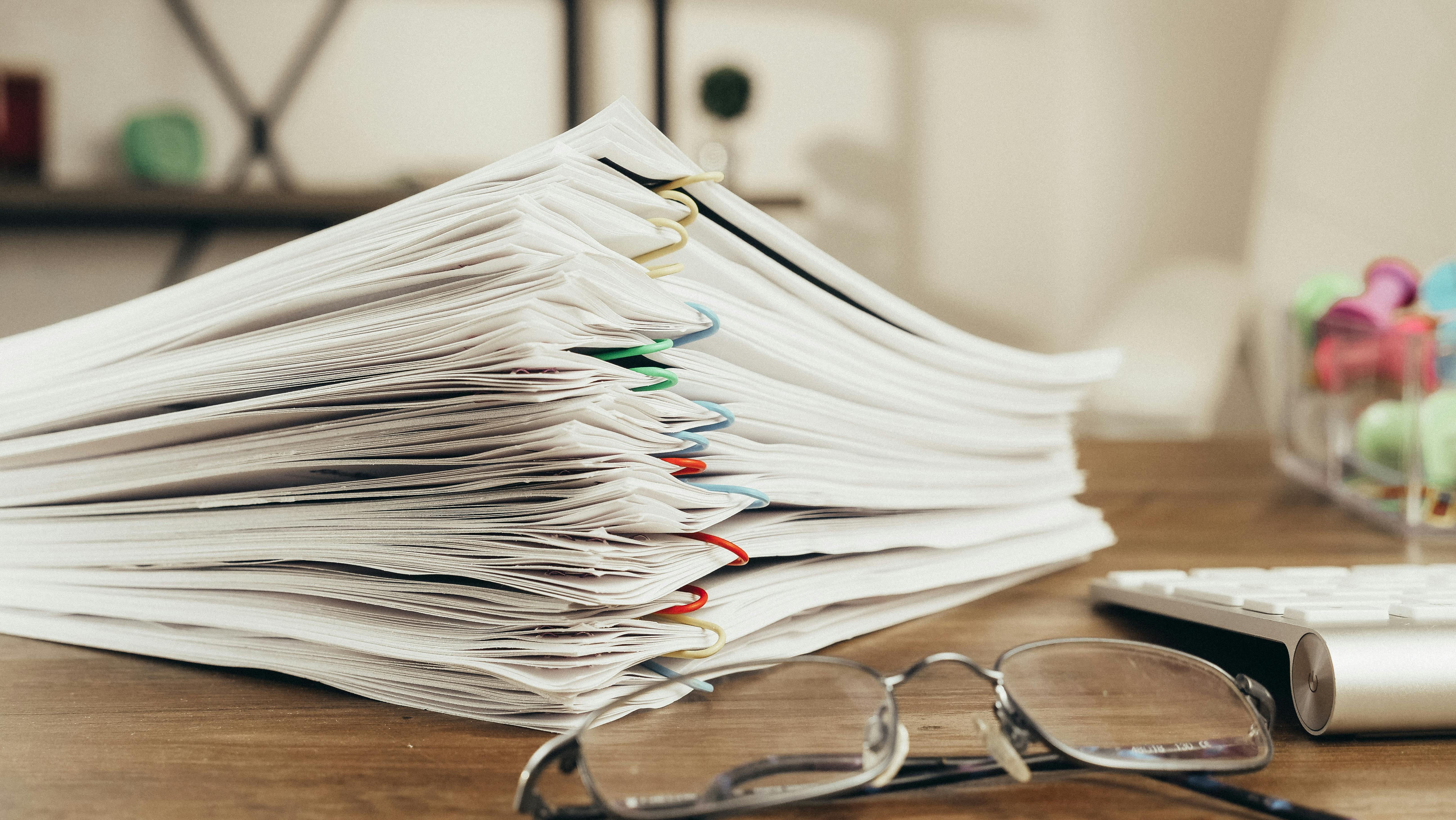 Stack of paper documents clipped together with colorful paper clips on a desk, with eyeglasses and a calculator nearby.