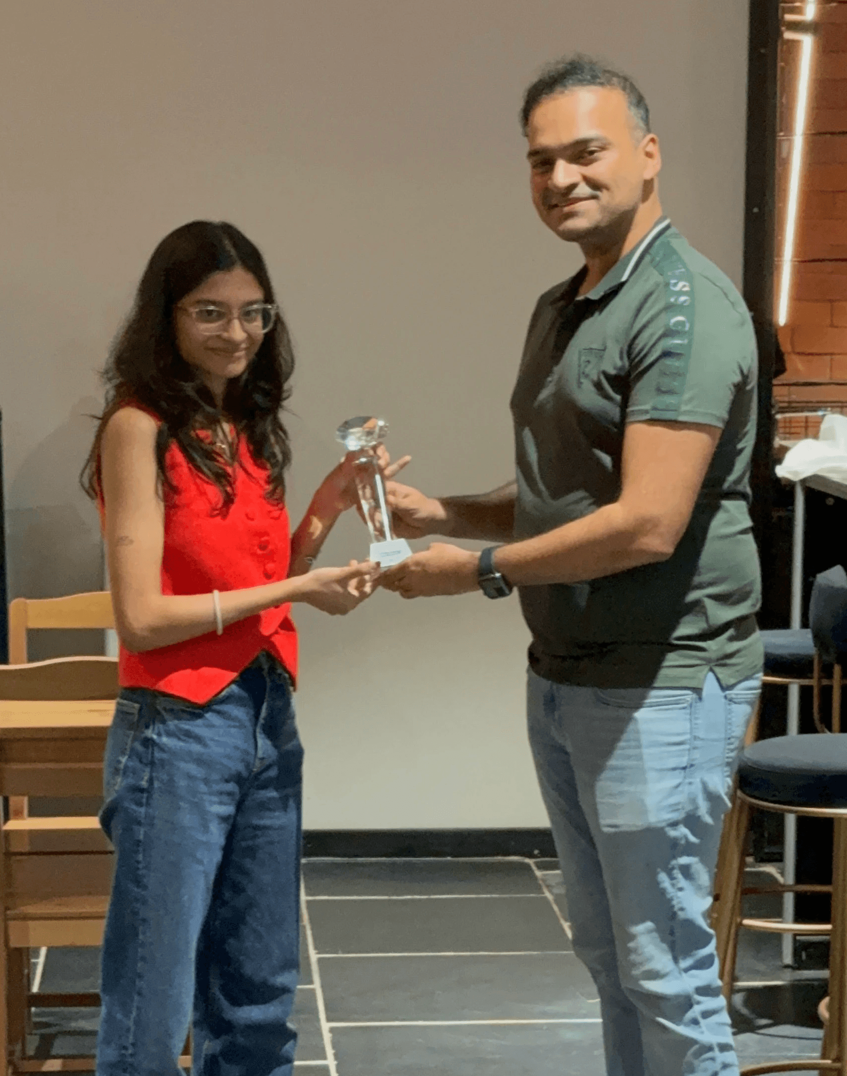 Una persona con una camiseta negra y pantalones de color claro está de pie en la cima rocosa de una montaña al atardecer, sosteniendo una cámara frente a su cara para tomar una foto. Un mar de nubes se extiende detrás de ellos, iluminado por la luz dorada del sol poniente.