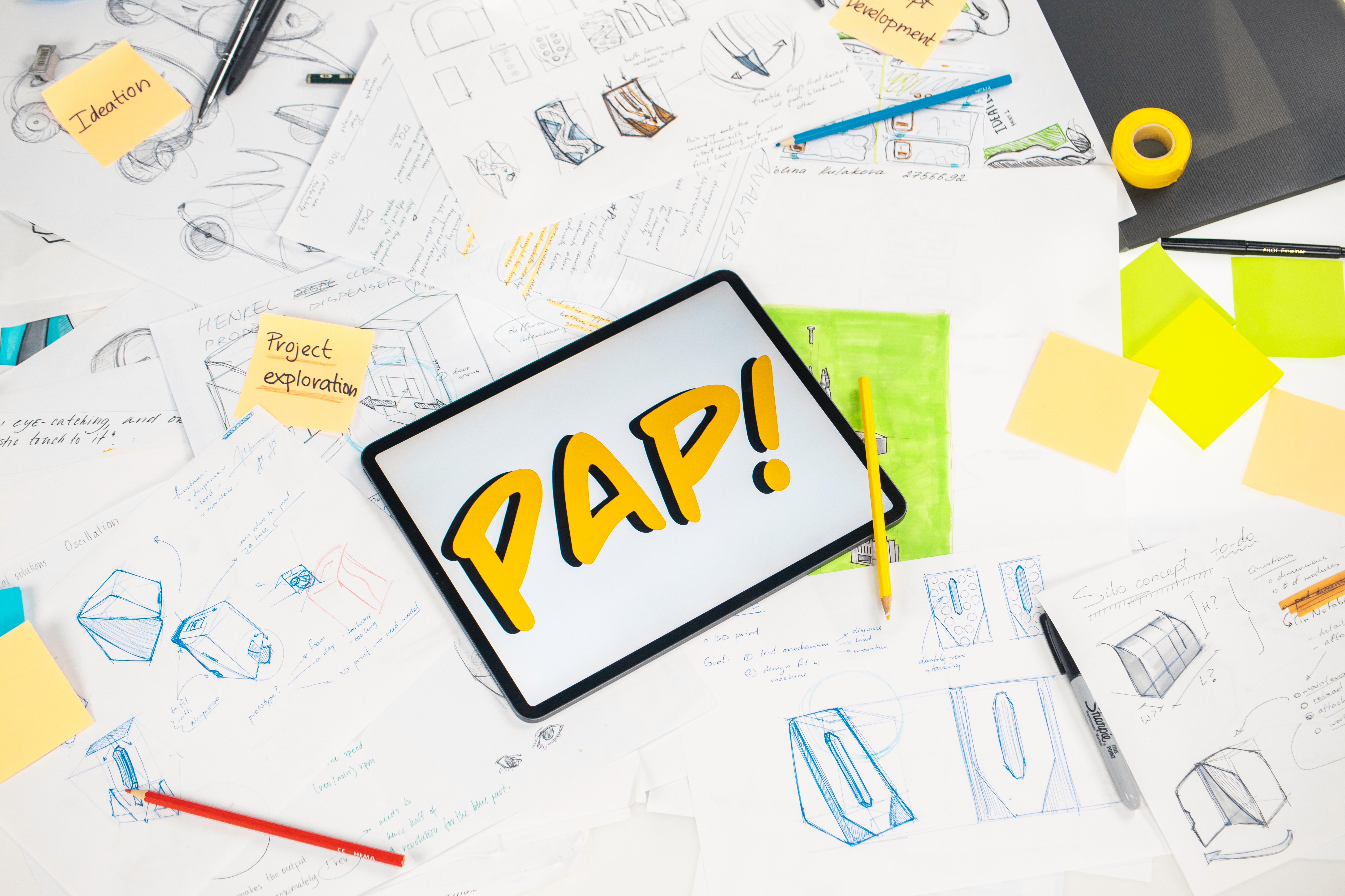 Tablet displaying "PAP!" on a desk covered with design sketches and notes labeled "Ideation" and "Project exploration." Creative, brainstorming mood.