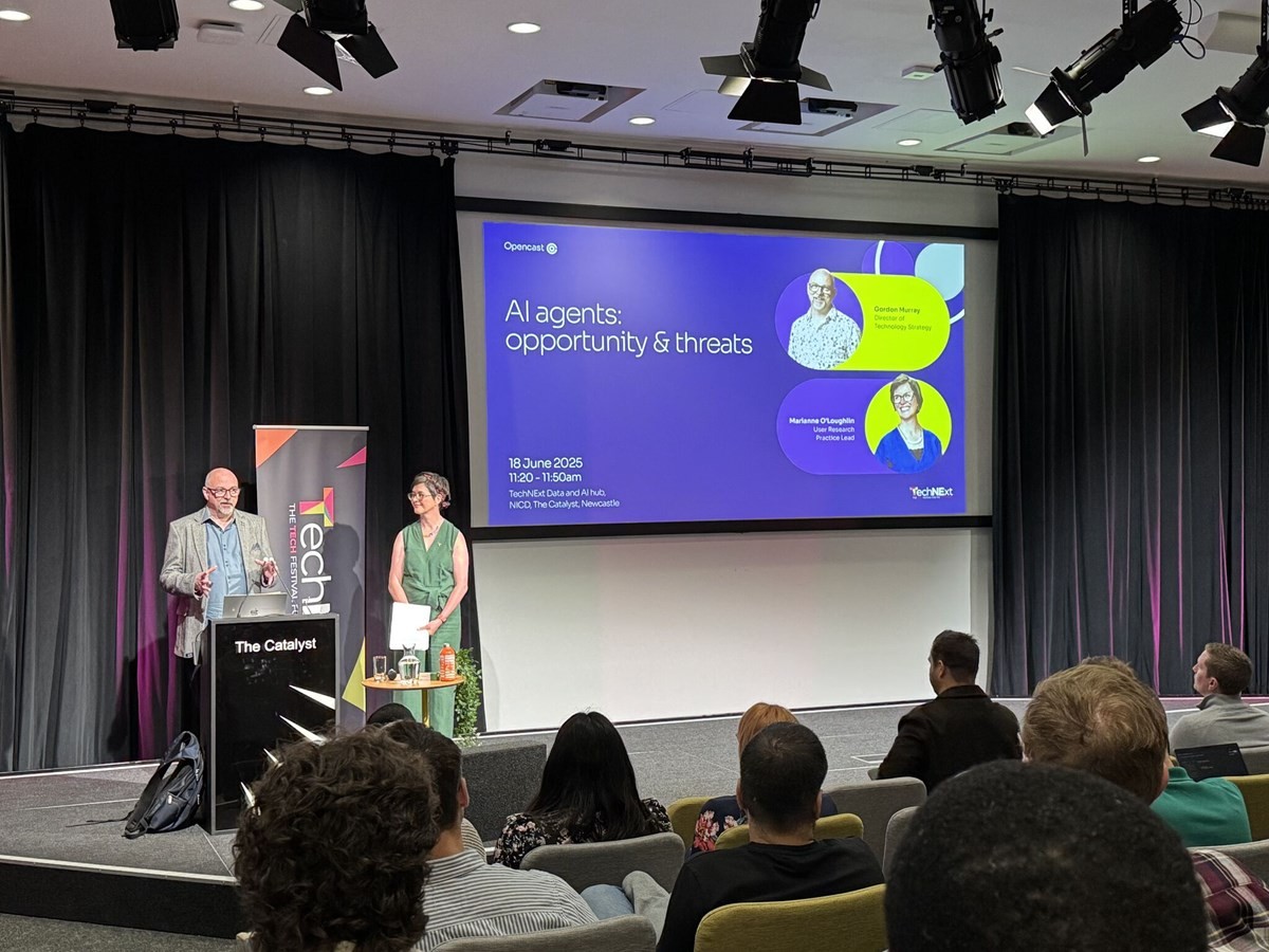 Two presenters standing on stage in front of a large screen displaying a slide titled ‘AI agents: opportunity & threats,’ with an audience seated and watching. The slide includes names, photos, and event details for a session at The Catalyst.