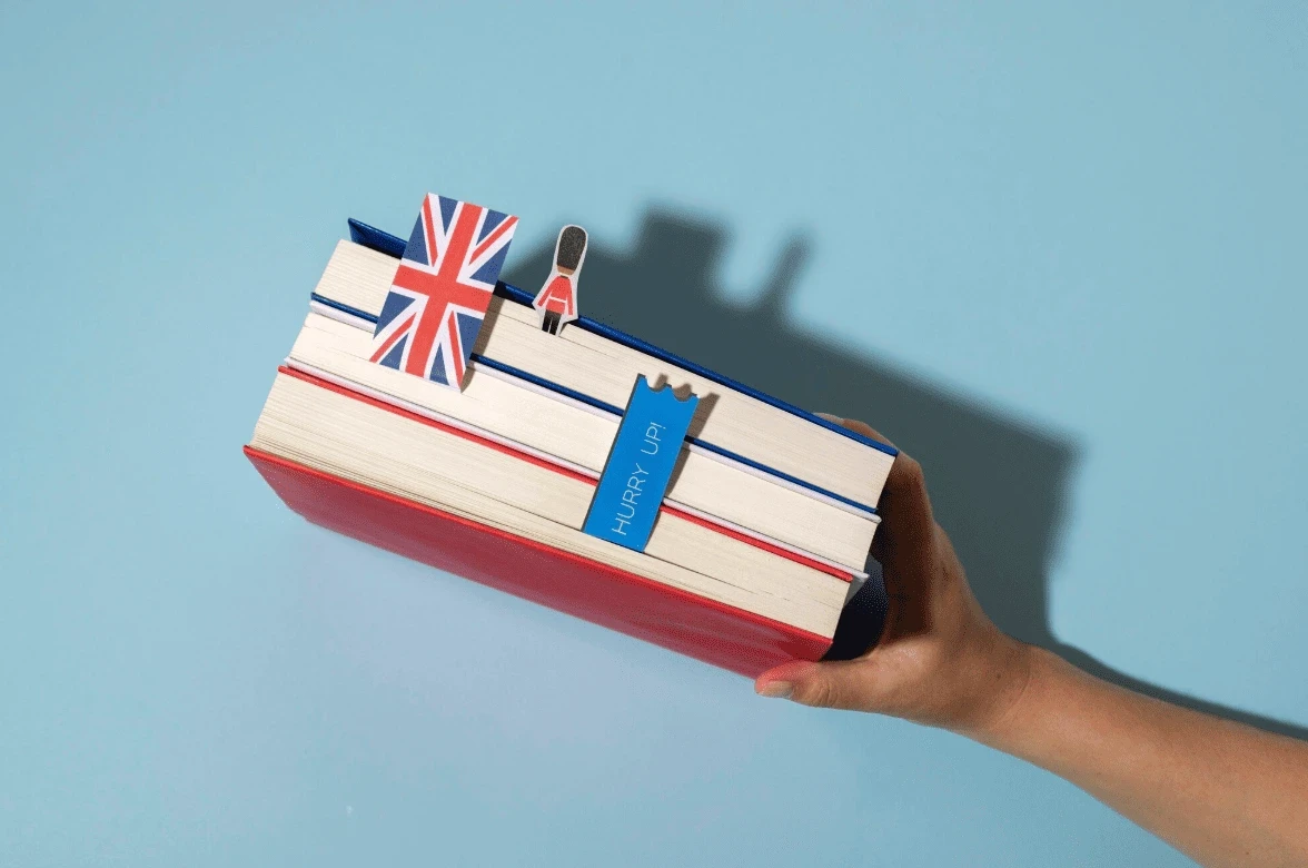 Hand holding a stack of books with British-themed bookmarks, including a Union Jack flag and a guard figurine, symbolizing English language learning or UK culture.
