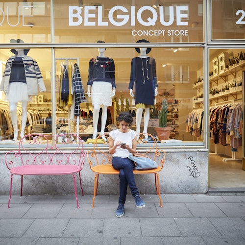 Orang duduk di bangku oranye di luar "Belgique Concept Store," dengan manekin dan pajangan pakaian di jendela.