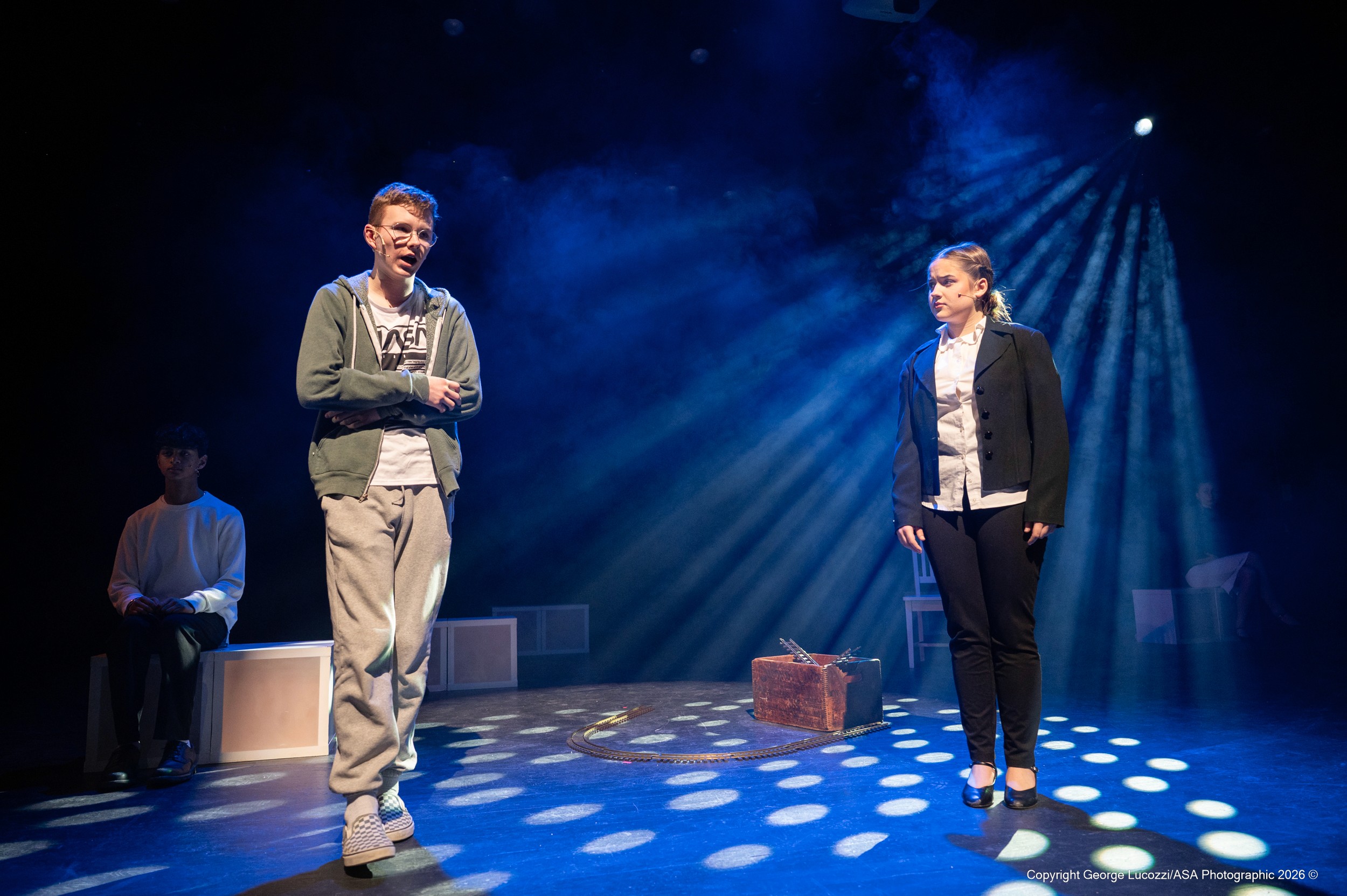 Christopher (left) and Reverend Peters (right) stand in dense haze, carved by angled blue-white gobo light that reveals strong aerial beams, supported by a cool top wash that frames their confrontation.