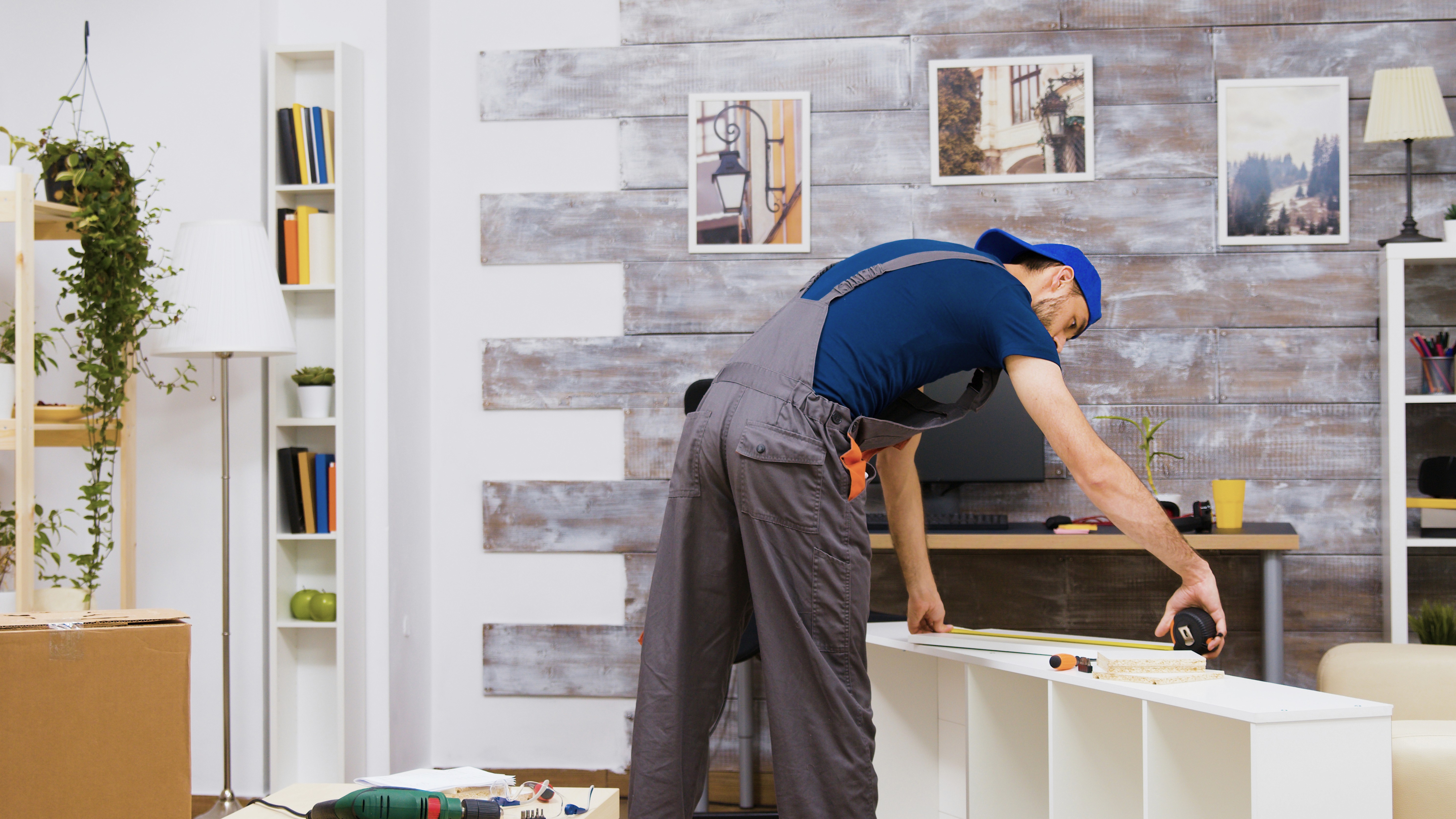 A person in overalls and a cap assembles white furniture in a modern living room for renovation. The setting is cozy, with plants, bookshelves, and rustic décor.