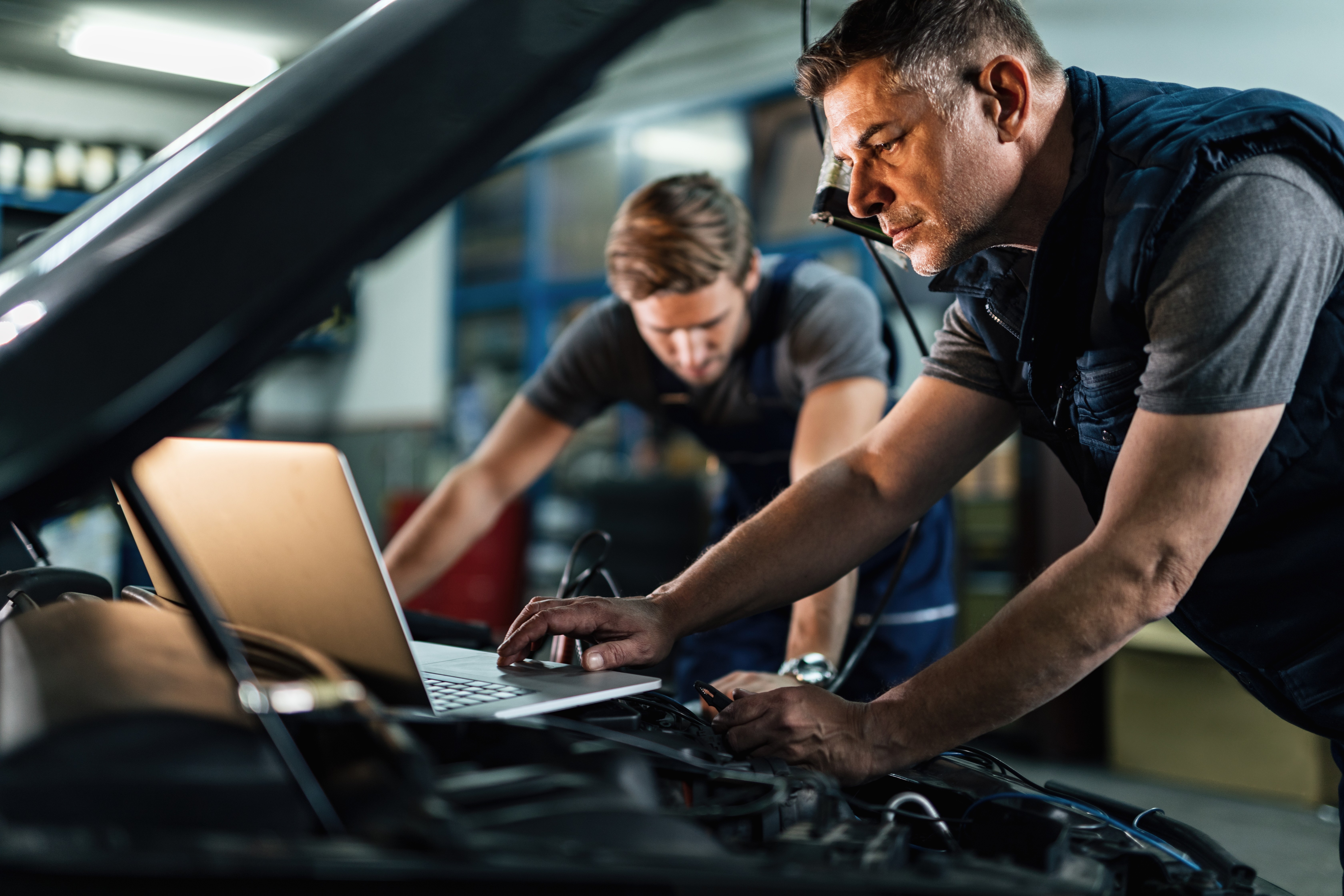 Two men in an auto repair shop preforming ADAS calibration on a vehicle with their laptop open