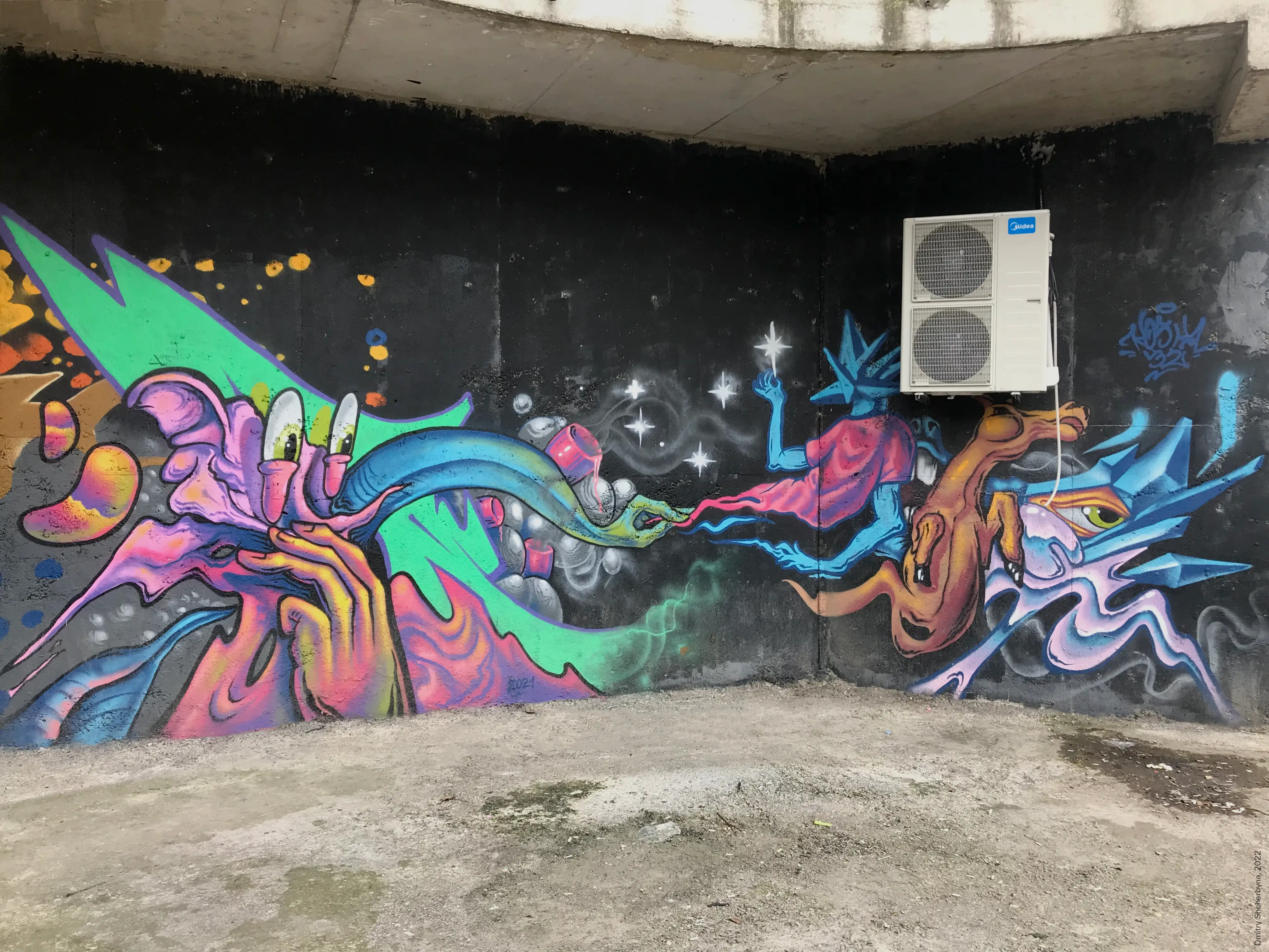 A mural on a dark wall beneath a concrete overhang depicts two fantastical creatures facing each other across a starry gap. On the left, there is a creature with neon green wings and pink and yellow flowing forms and bug-like eyes. On the right, there is another creature that is spiky and blue, with an orange body. The bright, psychedelic colors contrast with the black background.