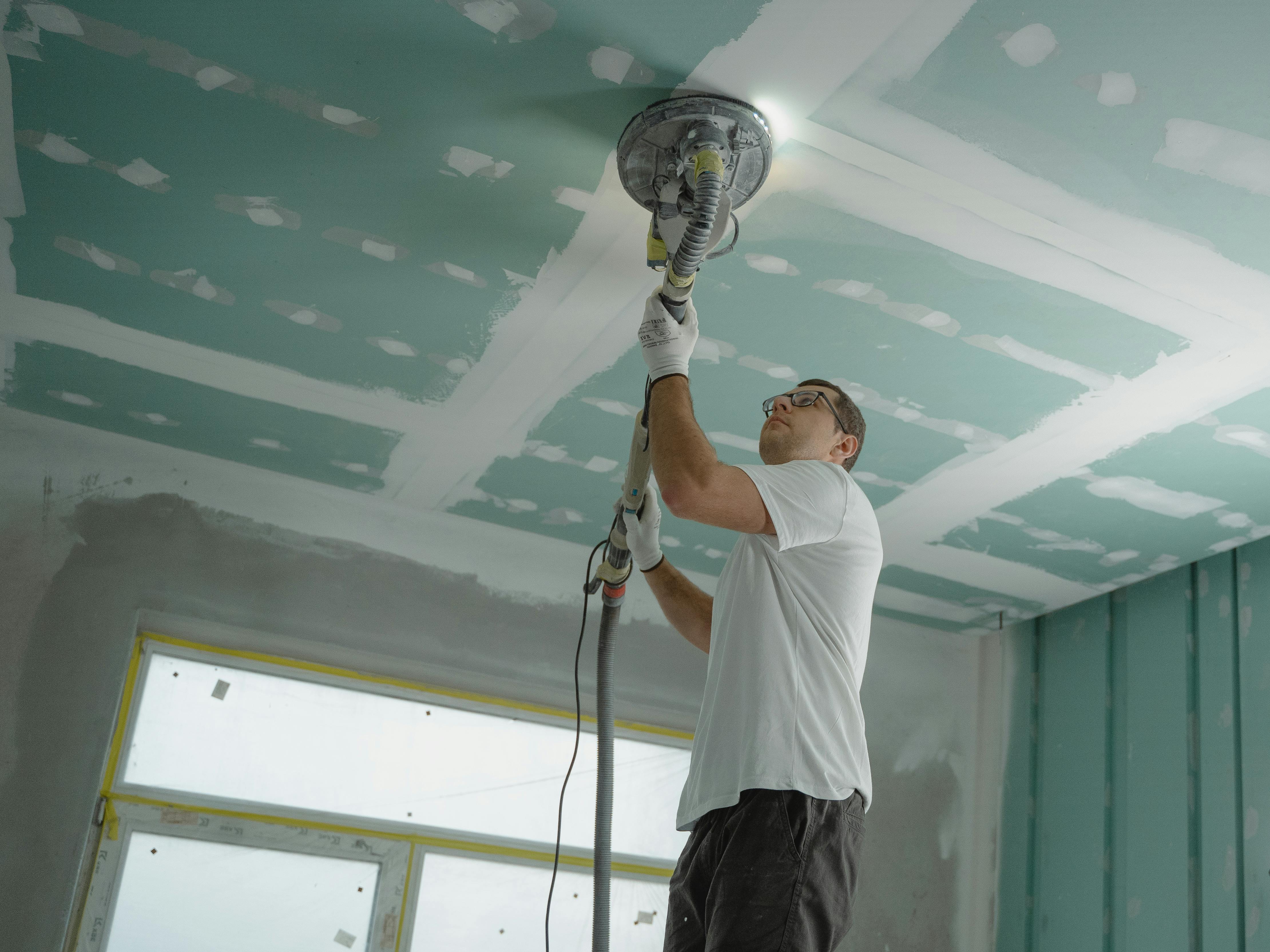 A person sanding a ceiling with a power sander in a room under renovation. They wear a white shirt, glasses, and gloves, focusing intently on their task.