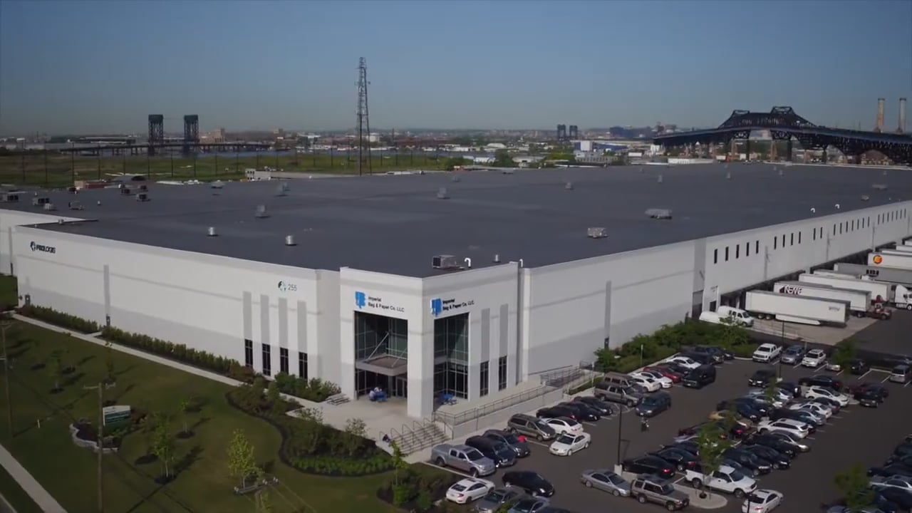 Aerial view of a large, modern warehouse facility with a gray roof, surrounded by a neatly landscaped parking lot filled with cars and trucks, set against a backdrop of industrial structures and a bridge under a clear blue sky.