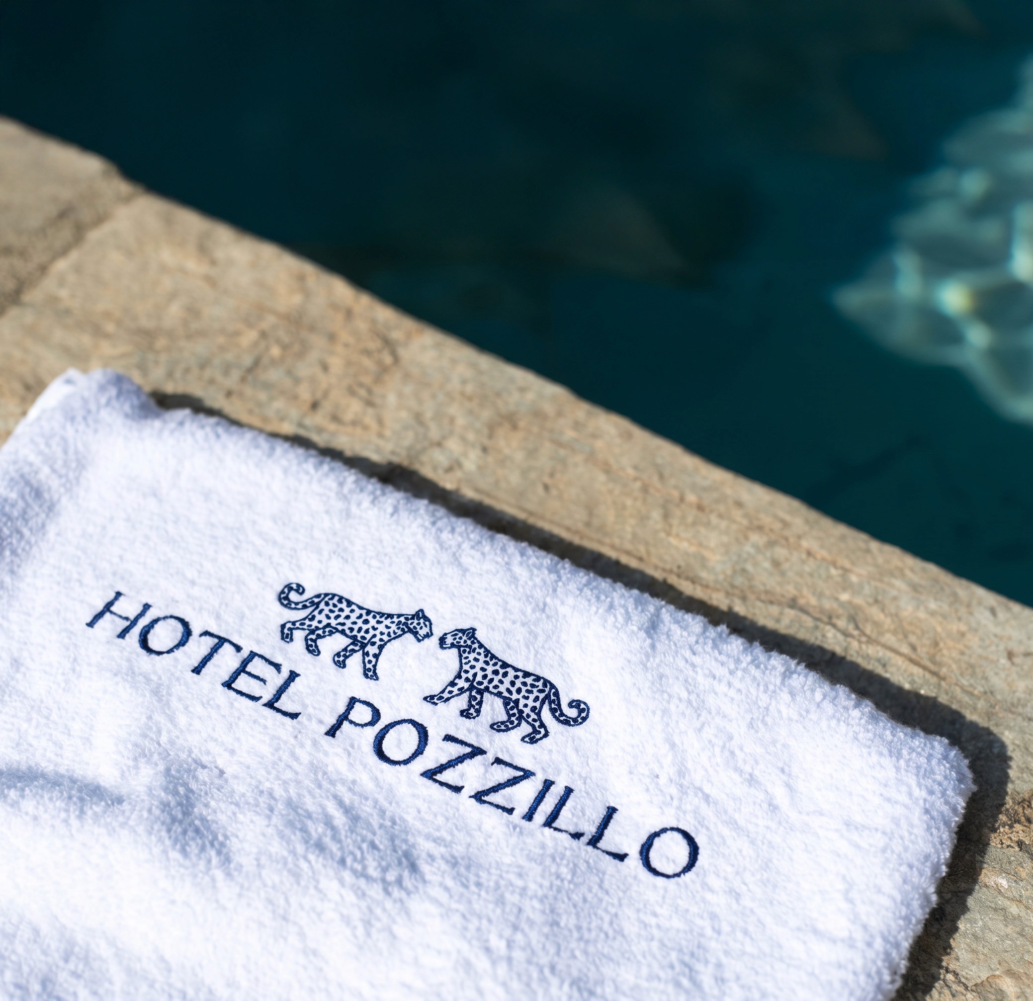 An white beach towel poolside with an embroided logo featuring two leopard in an elegant serif font in navy thread