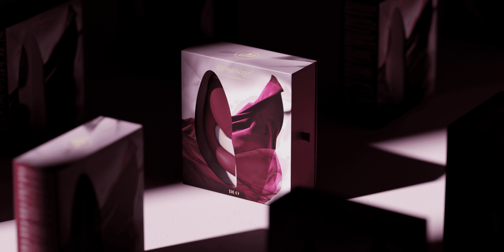 Markenwelt Womanizer „DUO" – Verpackung mit ovalem Sichtfenster und drapiertem Bordeauxstoff, inszeniert auf hellem Sockel in atmosphärischem Halbdunkel, mit goldenem „Womanizer The Original"-Schriftzug. EN: Womanizer „DUO" brand world – packaging with an oval die-cut window and draped burgundy fabric, staged on a light pedestal in atmospheric low light, with the gold „Womanizer The Original" wordmark.