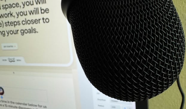 Close-up image of a black microphone positioned in front of a computer screen displaying text. The microphone's mesh detail is prominent, suggesting a setting for recording audio or podcasting. The blurred background screen features text related to goals or work, hinting at professional or creative workspace environment.