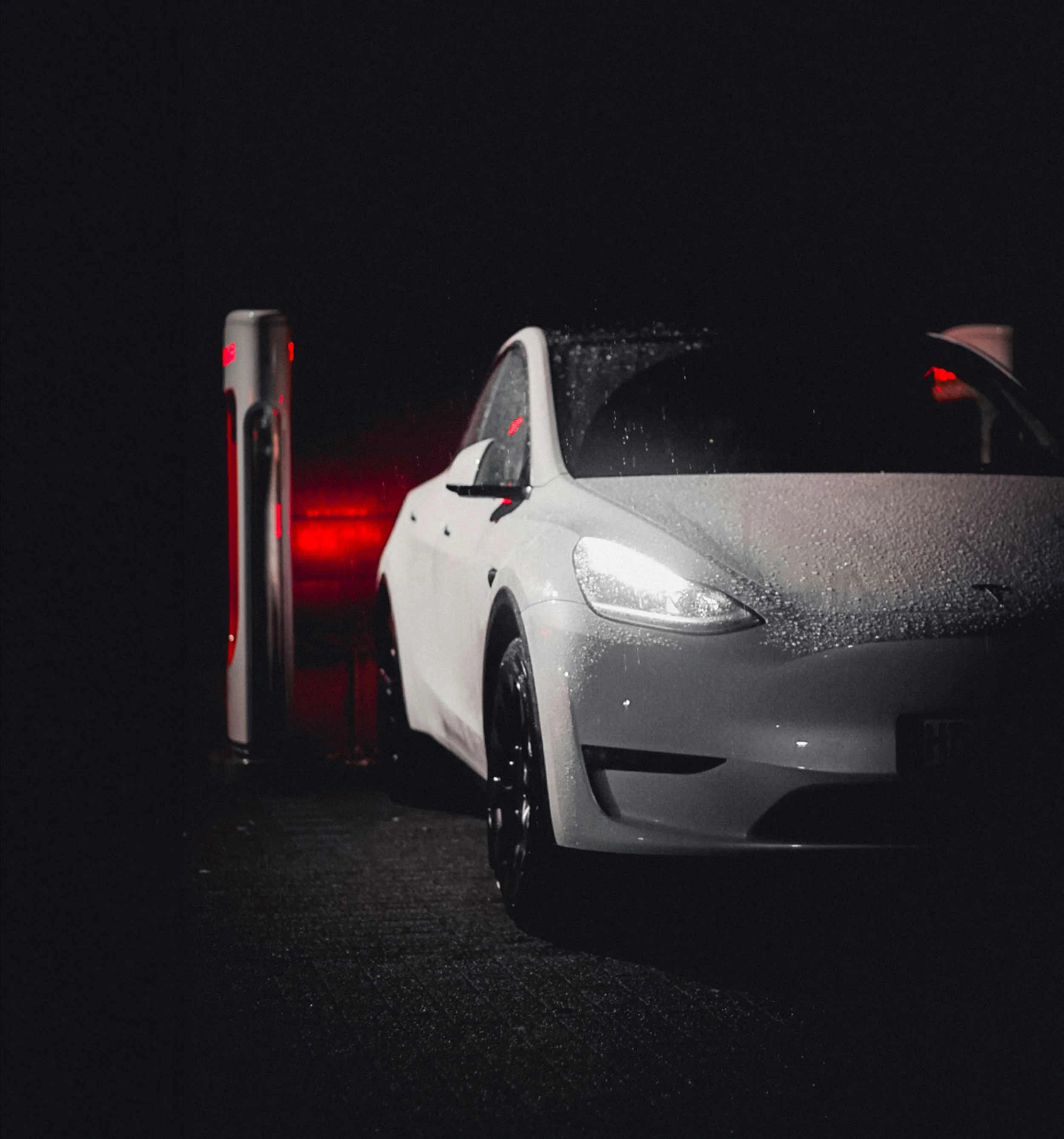 En vit elbil laddar vid en laddstation i mörker med regndroppar på bilen och rött ljus från laddaren i bakgrunden.