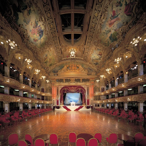 Ornate ballroom with a large dance floor, chandeliers, detailed ceiling artwork, red velvet curtains on stage, and red seating.