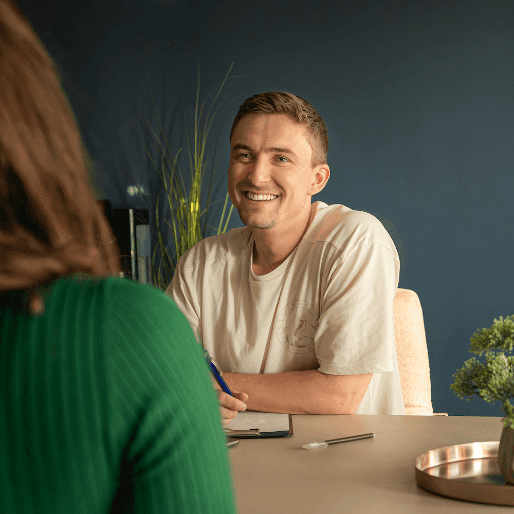 Zwei Personen sitzen sich an einem Tisch gegenüber, eine trägt ein grünes Oberteil, die andere ein helles T-Shirt und hält einen Stift über einem Klemmbrett