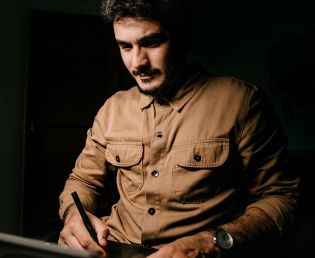 Man sitting in a dimly lit room, wearing a beige shirt and looking contemplative.