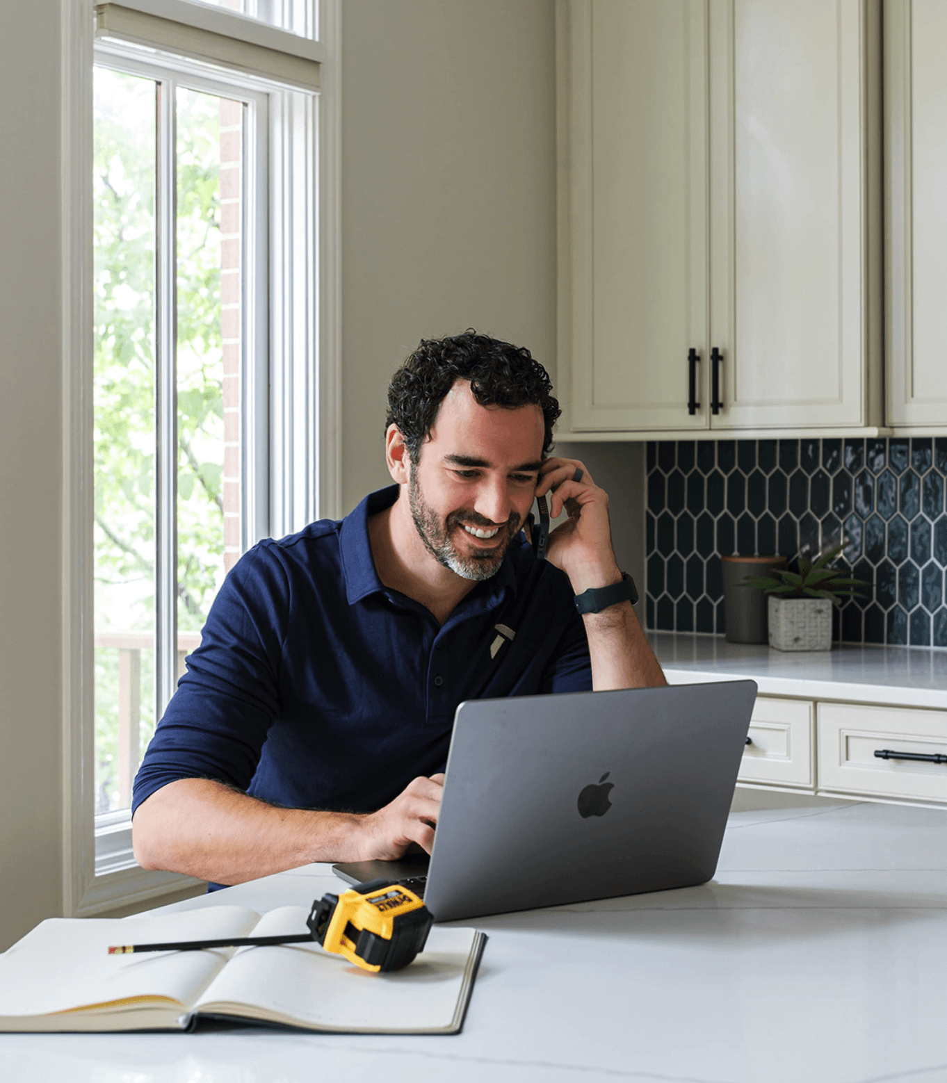 a man sits at a kitchen island, he's on a cell phone and looking at a laptop, a notebook and measuring tape are on the counter next to him