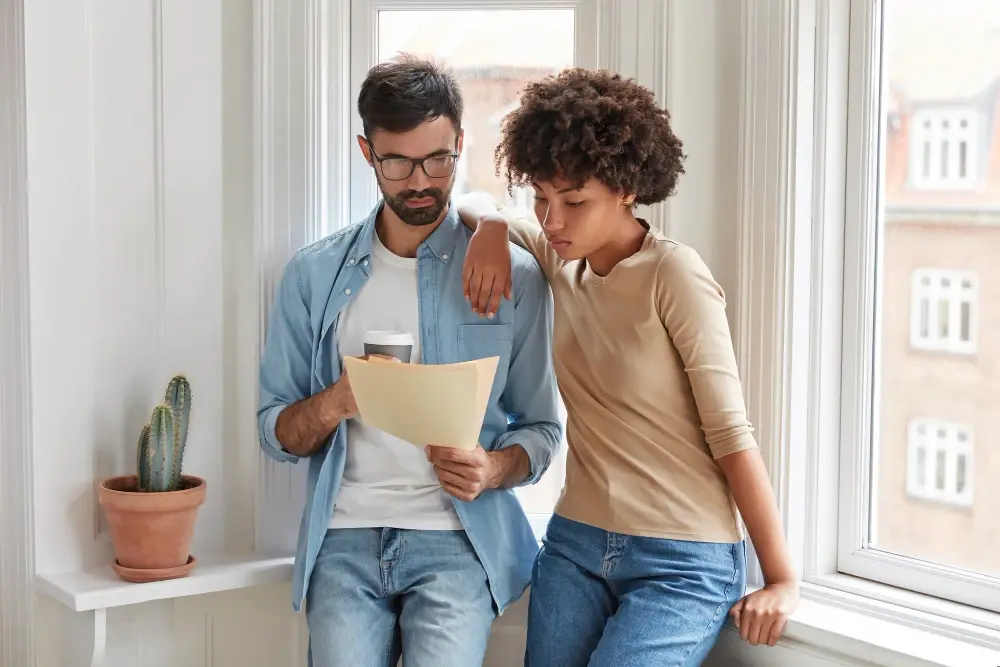 A couple stands by a window reviewing financial documents together, both appearing thoughtful and focused. The image represents the challenges and decisions involved in financing investment properties with Chris Lewis Home Loans.
