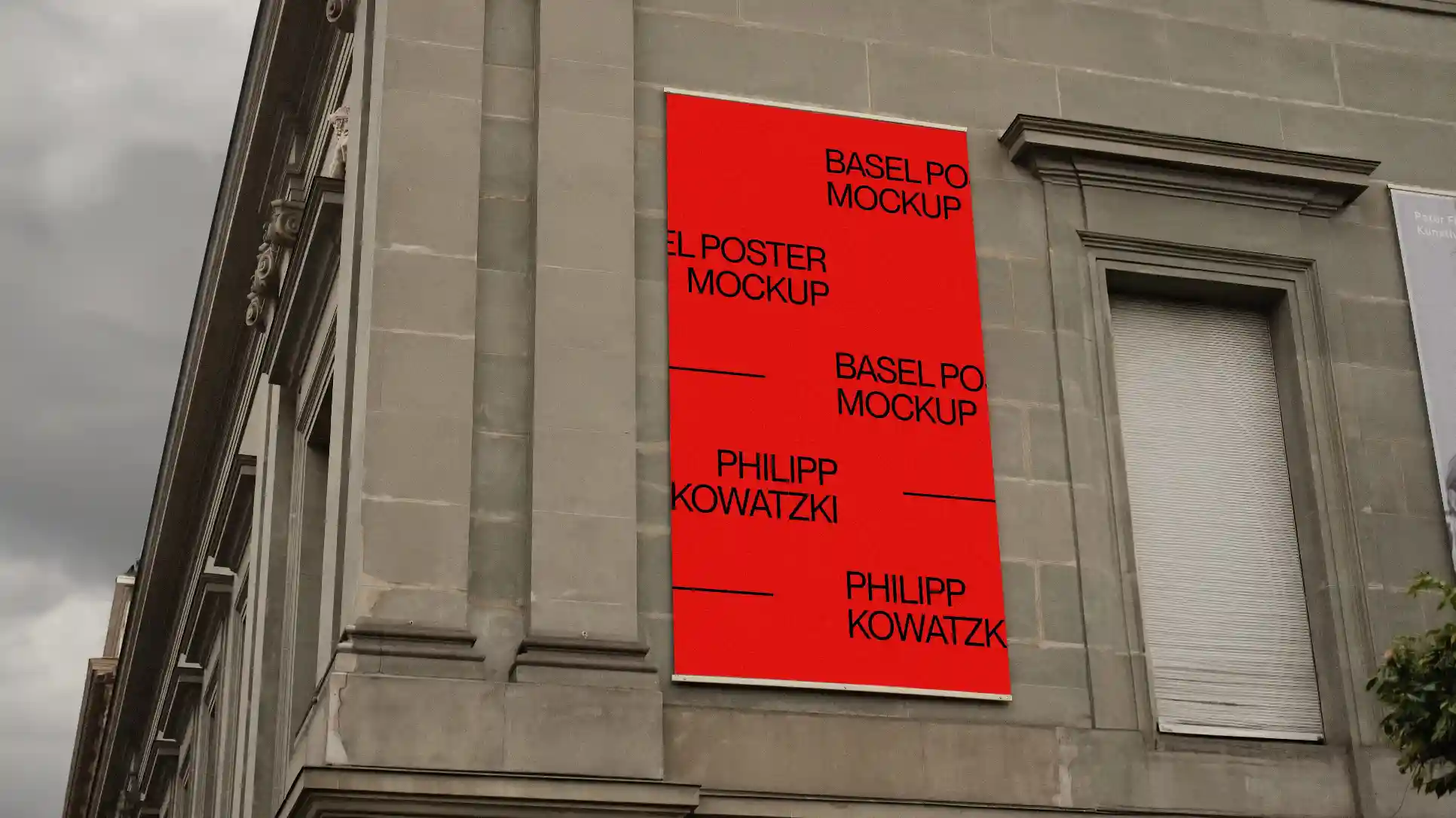 A vertical poster mockup displayed on the facade of a classical stone building. The bold red poster contrasts against the gray architecture, surrounded by ornate detailing, under an overcast sky.