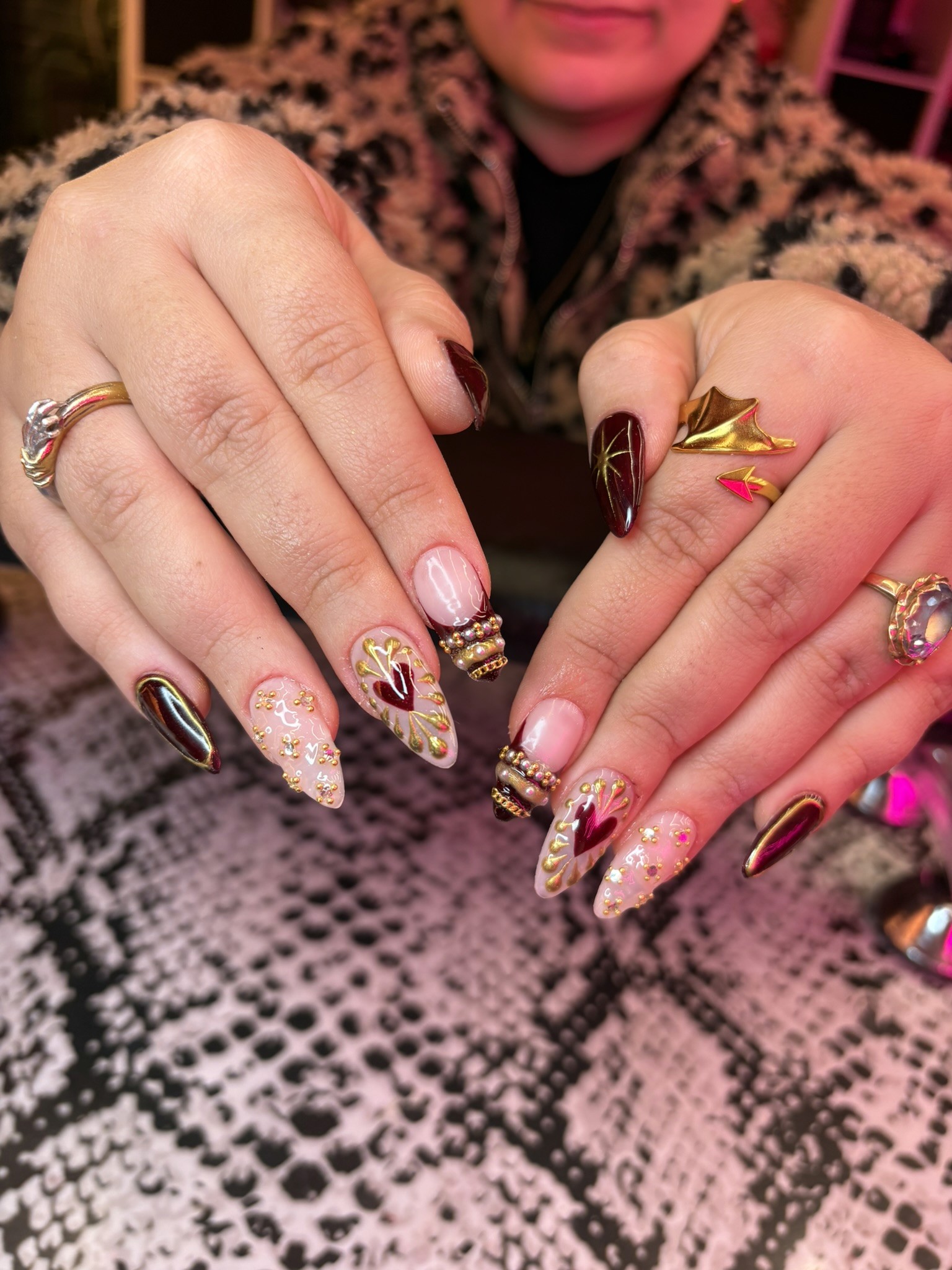 Almond and stiletto-shaped nails with a base of deep burgundy and sheer nude. Features hand-painted gold filigree surrounding blood-red hearts, detailed 3D gold and pearl "crown" accents at the cuticles, and gold-flecked glitter.