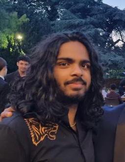 Woman in sunglasses and a brown coat stands against a vibrant blue wall. Sunlight casts a dramatic shadow, creating a confident, stylish atmosphere.