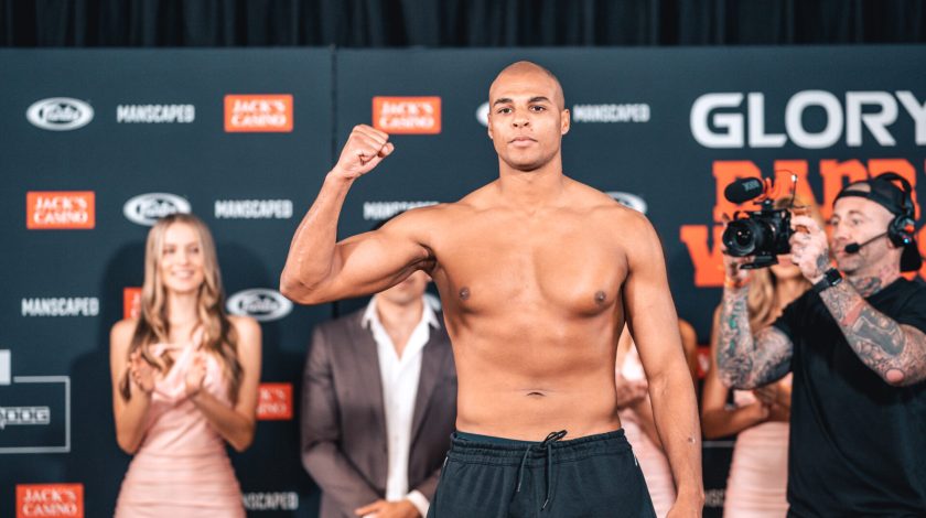 Levi Rigters confidently poses shirtless with a raised fist during a weigh-in event, surrounded by people and photographers, against a branded backdrop featuring logos for "Glory" and "Jack's Casino."
