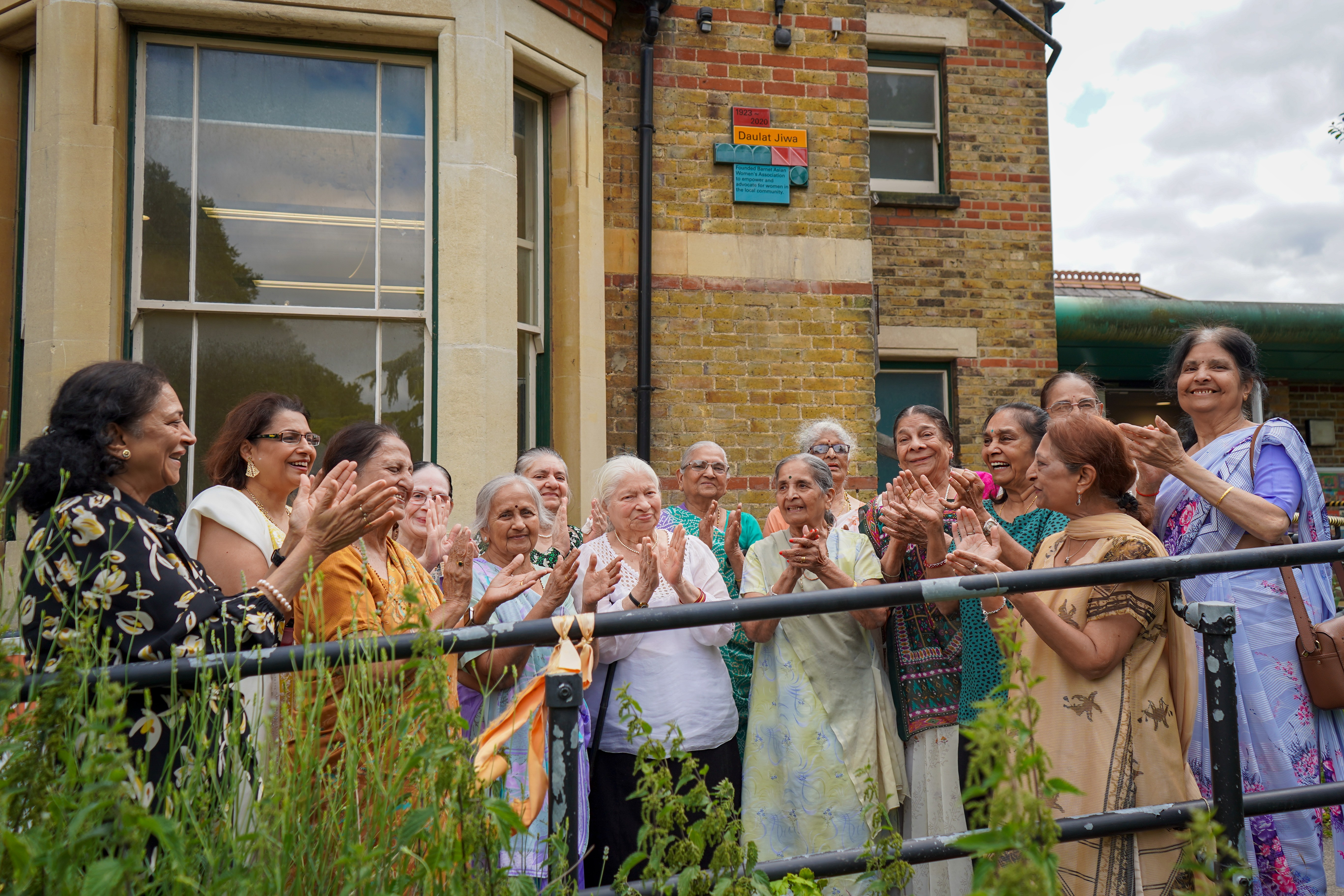 Group of women standing and clapping in front of a building near the Barnet Legend plaque for Daulat Jiwa
