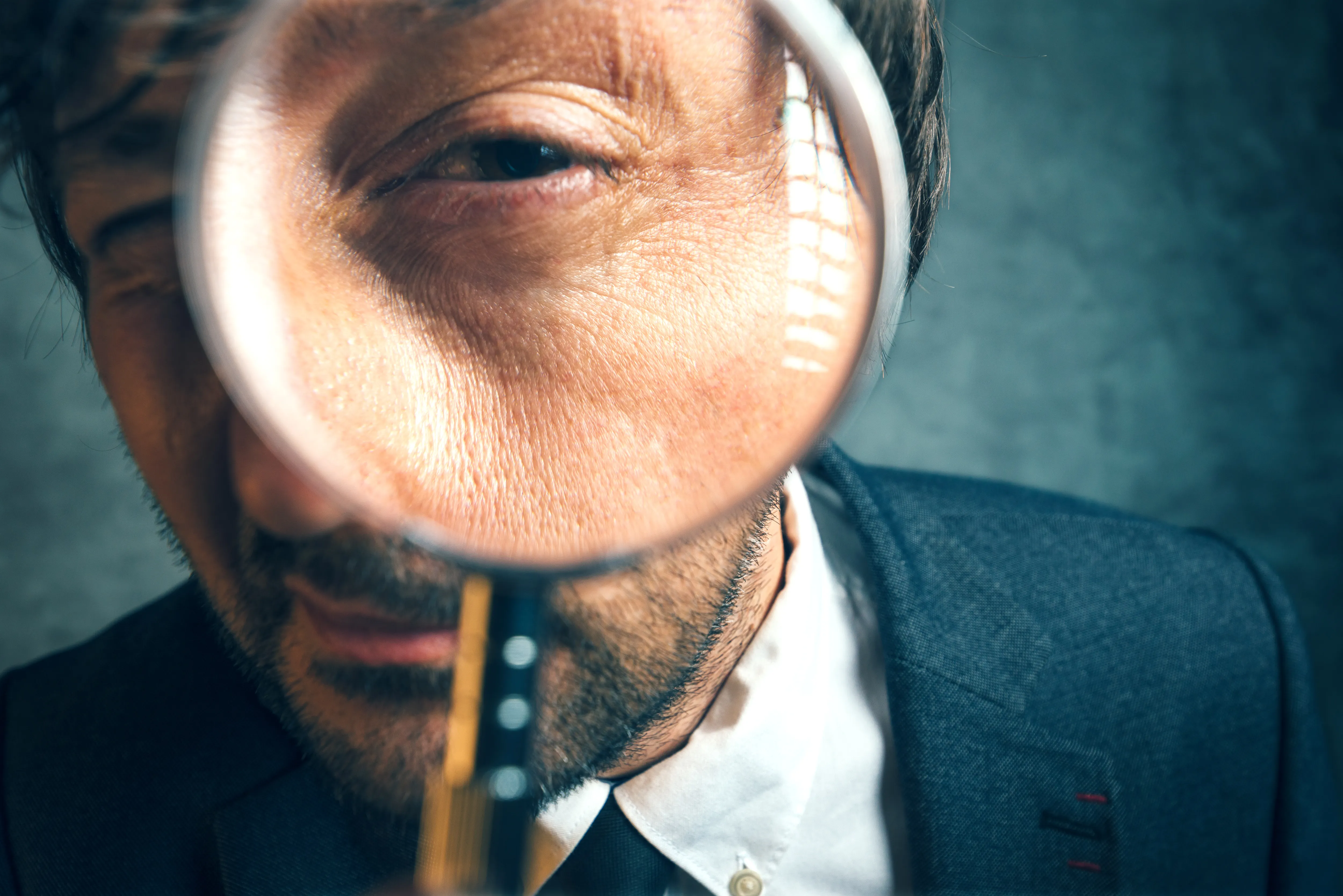 Close-up of a man in a suit looking through a magnifying glass, enlarging part of his face.