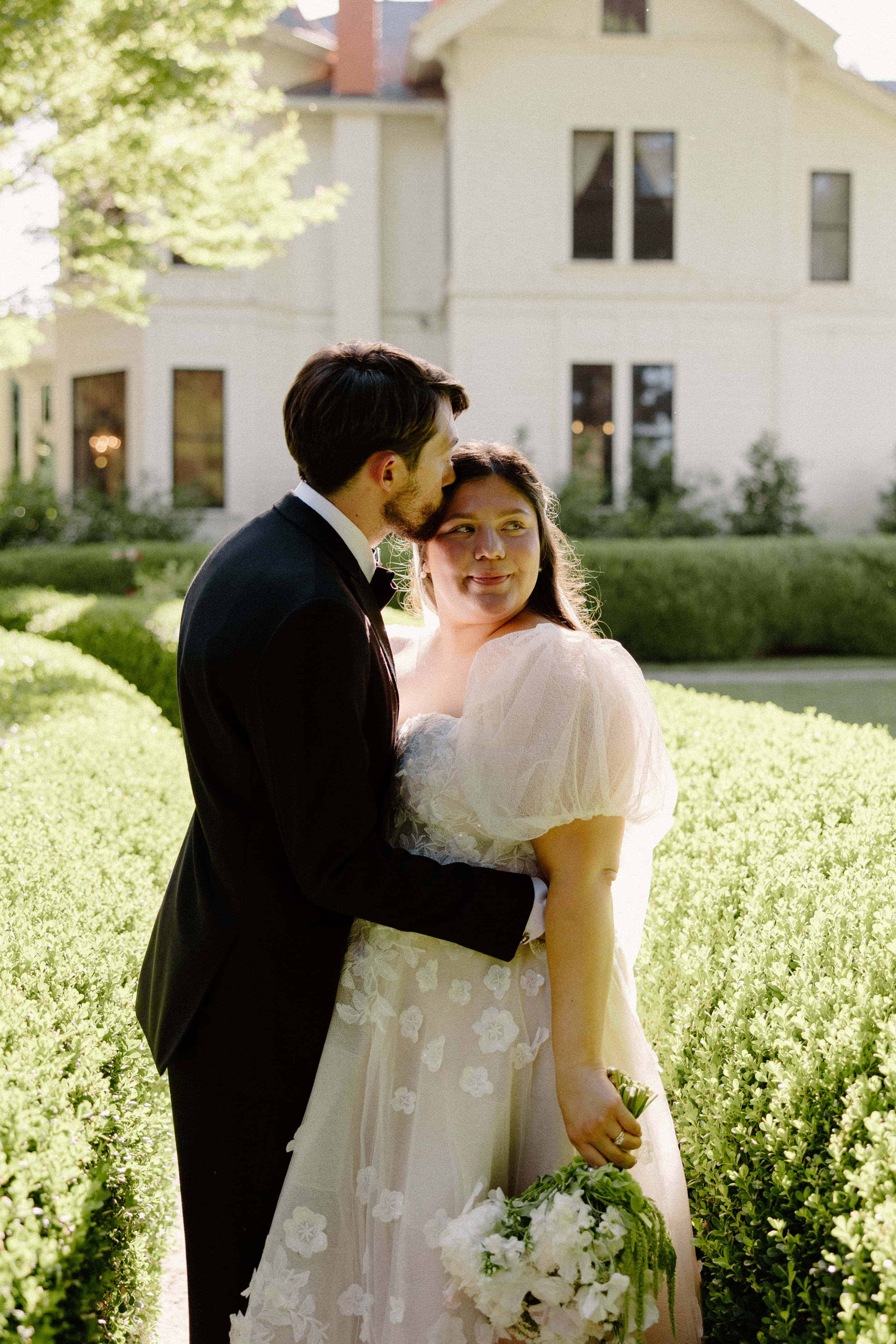A man in a black suit kisses a woman on the cheek. The woman is wearing a white floral dress and holding a bouquet. They are standing in a garden in front of a white building.