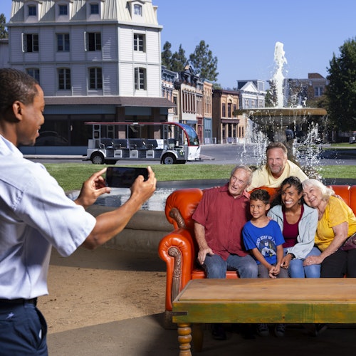 A man takes a photo of five people seated on an orange couch in front of a fountain with buildings in the background.