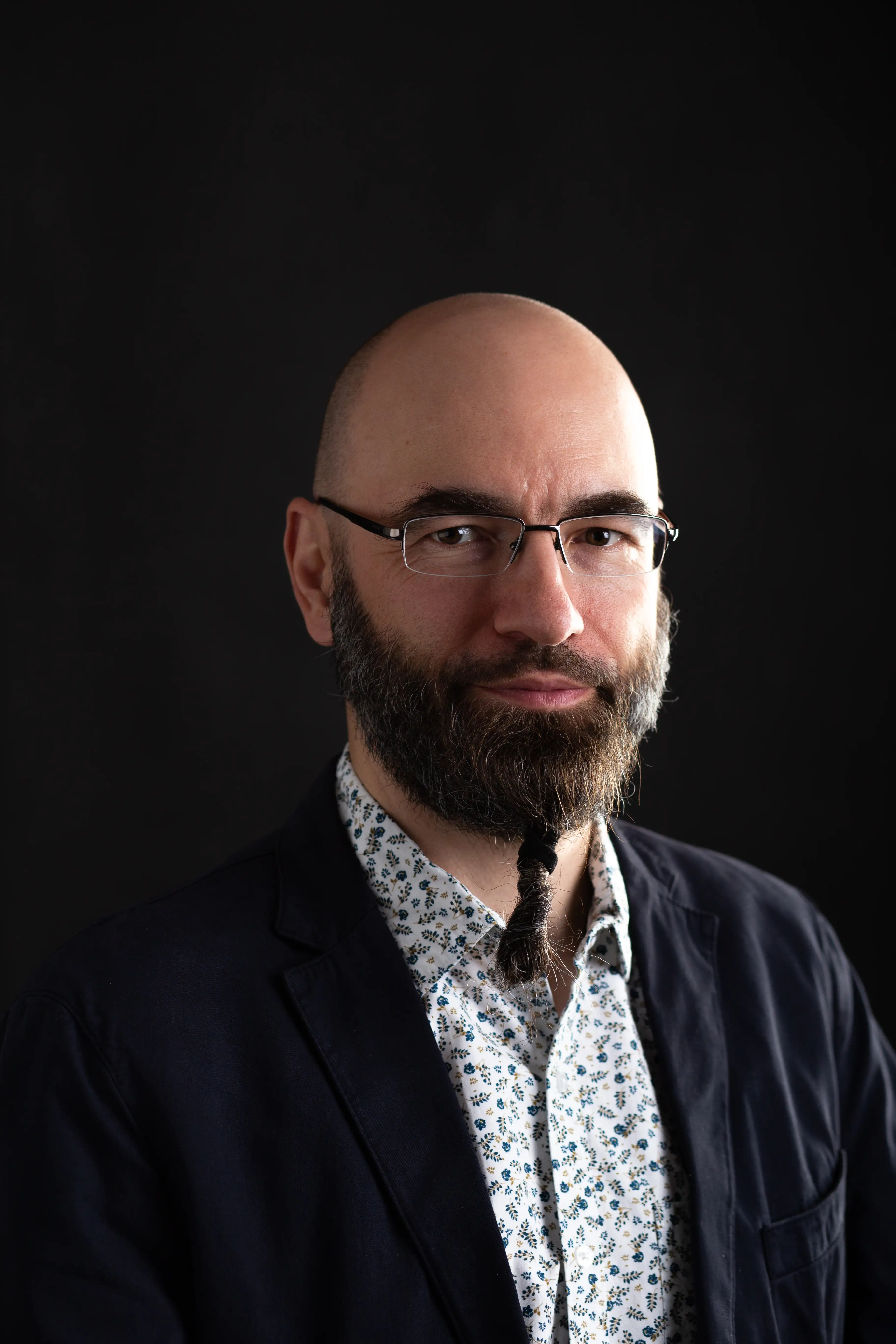 Mężczyzna z łysą głową, okularami i gęstą brodą z warkoczem na brodzie, ubrany w kwiecistą koszulę i ciemną marynarkę, pozuje na prostym czarnym tle.