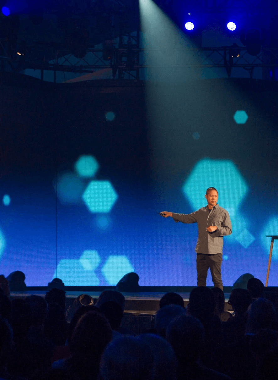 Speaker presenting on stage at C2 conference, addressing an audience under blue stage lighting.