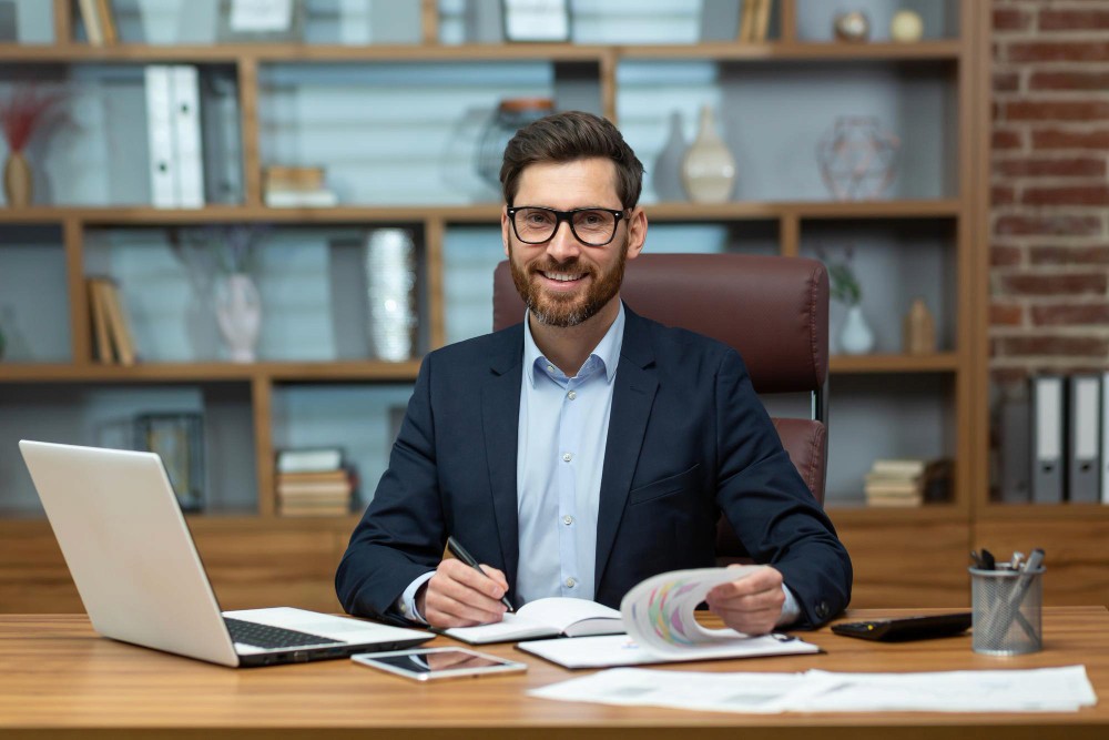 A smiling man with glasses and a beard sits at a desk in an office, wearing a suit jacket and holding a pen and documents.