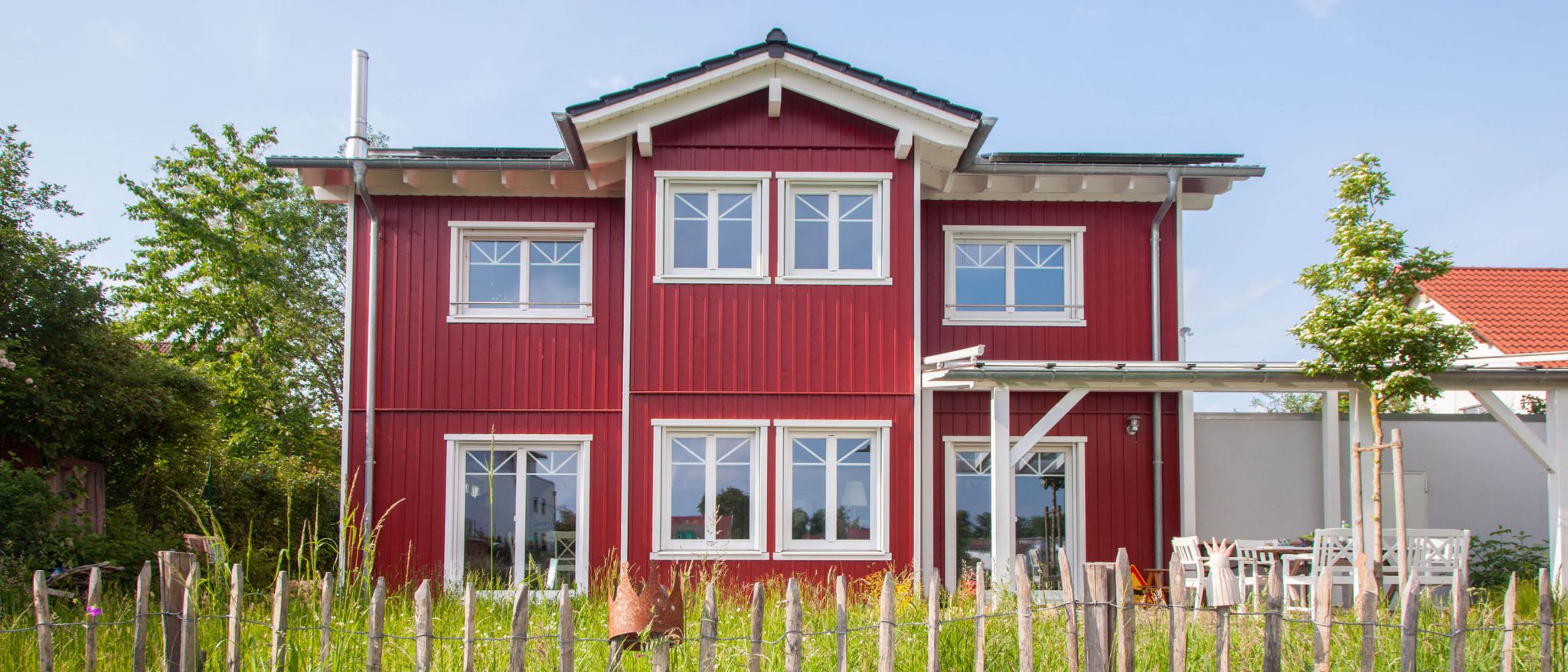 Rotes Sommerhaus mit weißen Sprossenfenstern, Holzverkleidung und einem Metallschornstein.